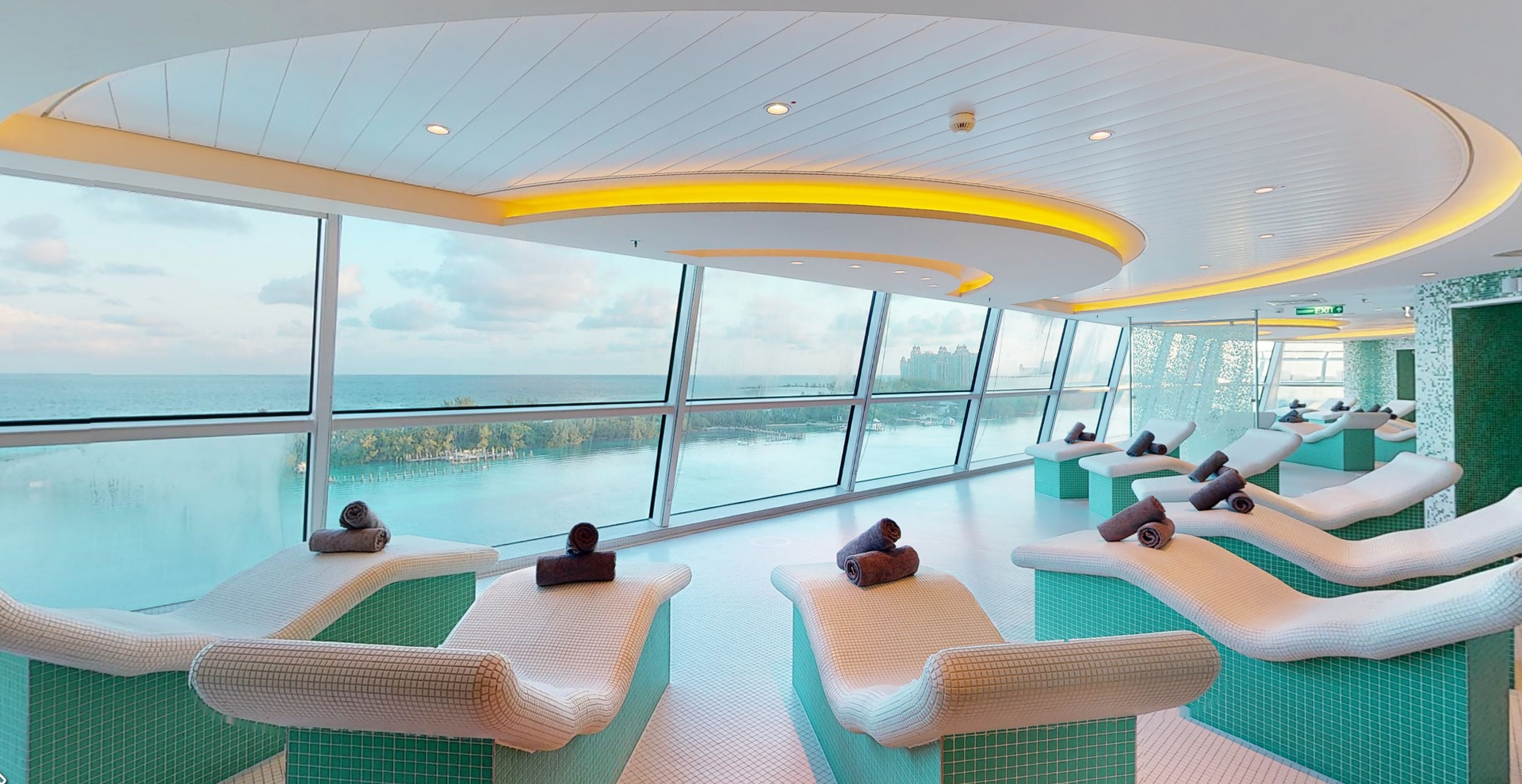 A relaxation room with lounge chairs and a view of the ocean through large windows; turquoise and white decor.