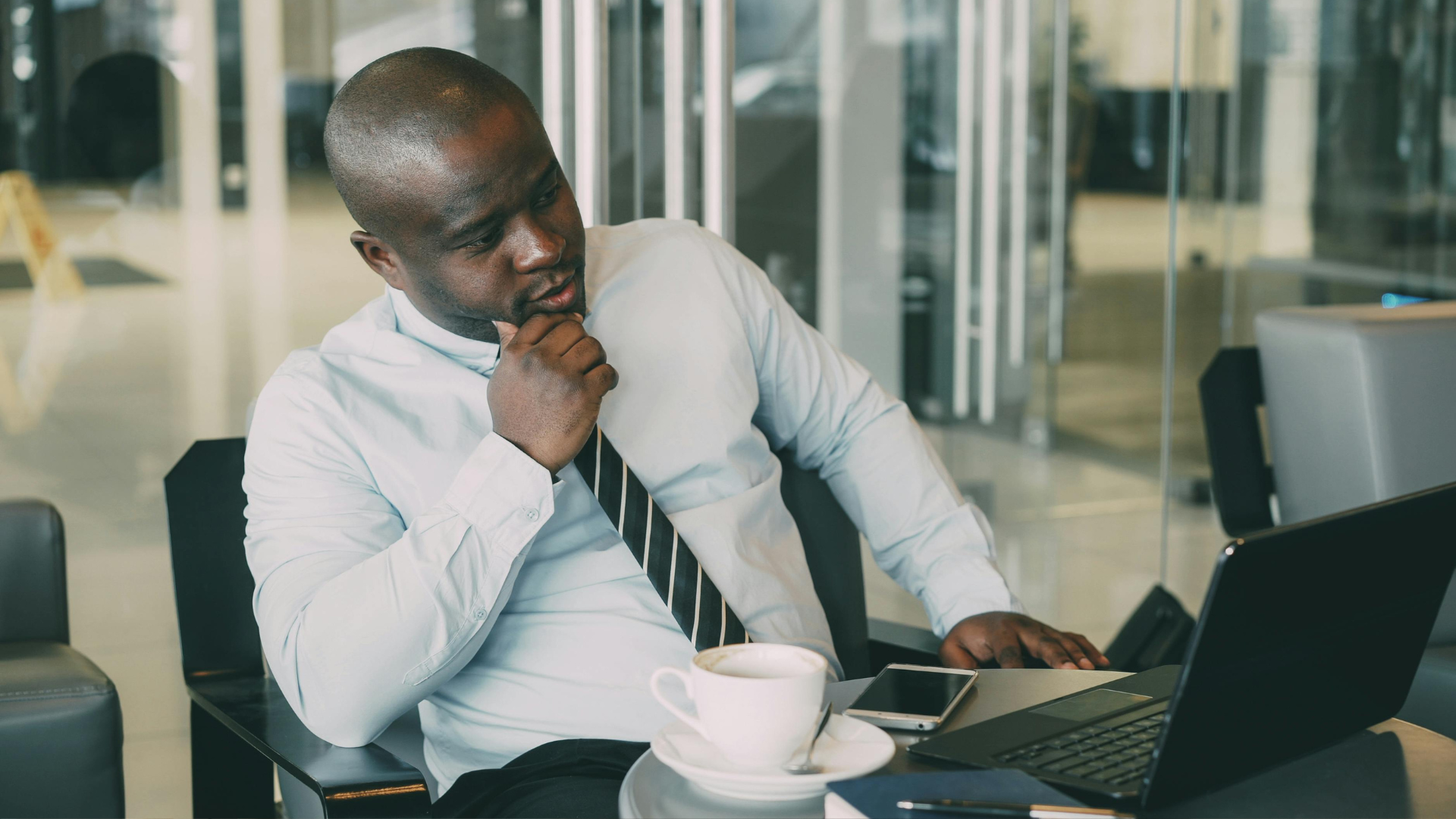 Man in a white shirt thinking at a laptop with a coffee cup on a desk in an office.