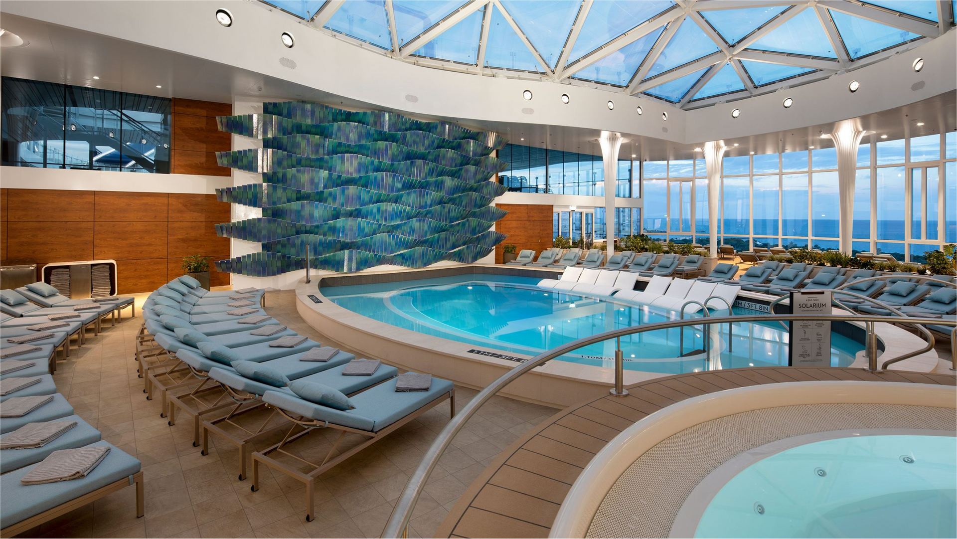 Indoor pool area with a pool, hot tub, and lounge chairs under a glass ceiling, with a blue water wall feature.