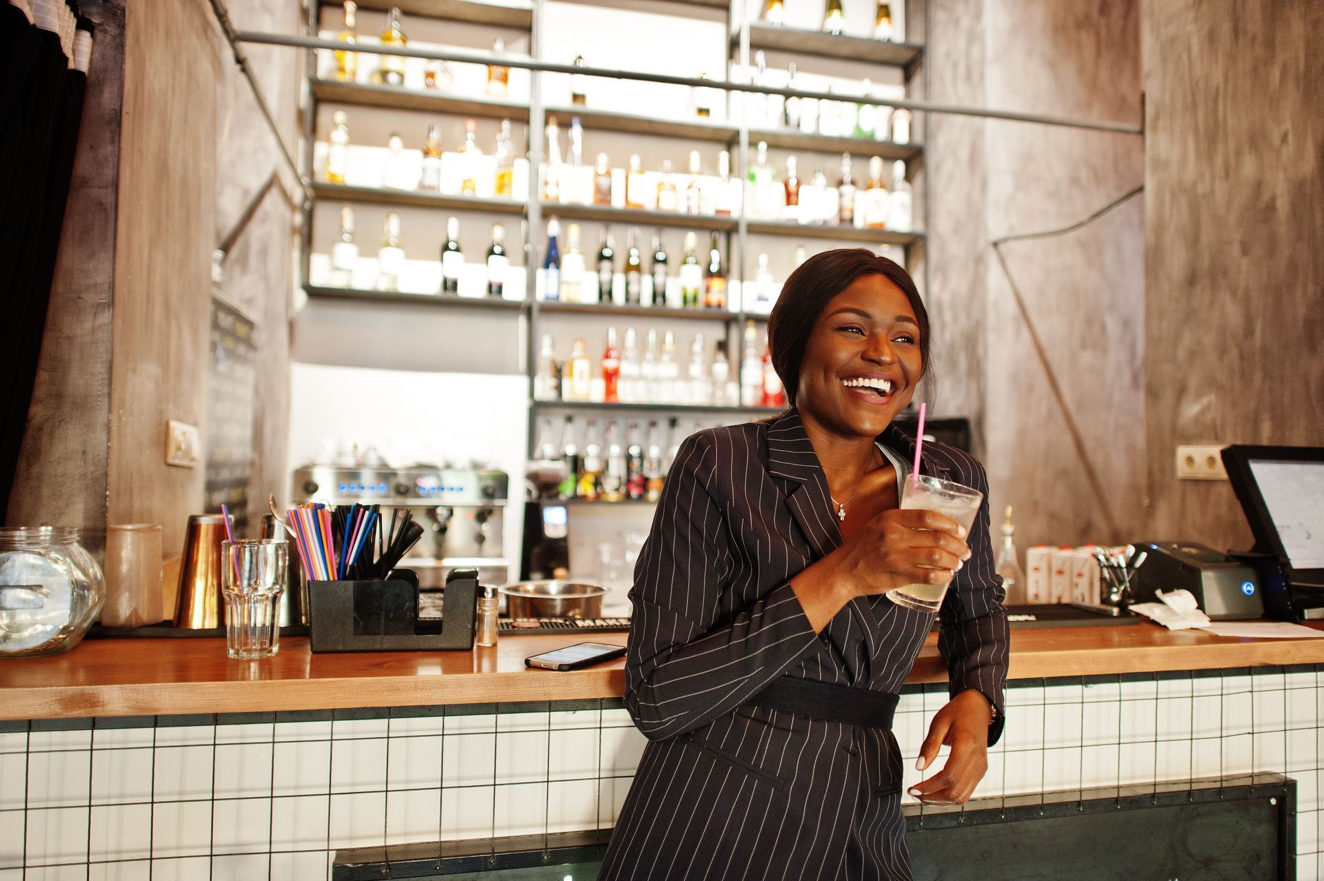 A smiling Black woman in a suit leans on a bar, holding a drink with a straw. Bar with shelves of bottles is in the background.