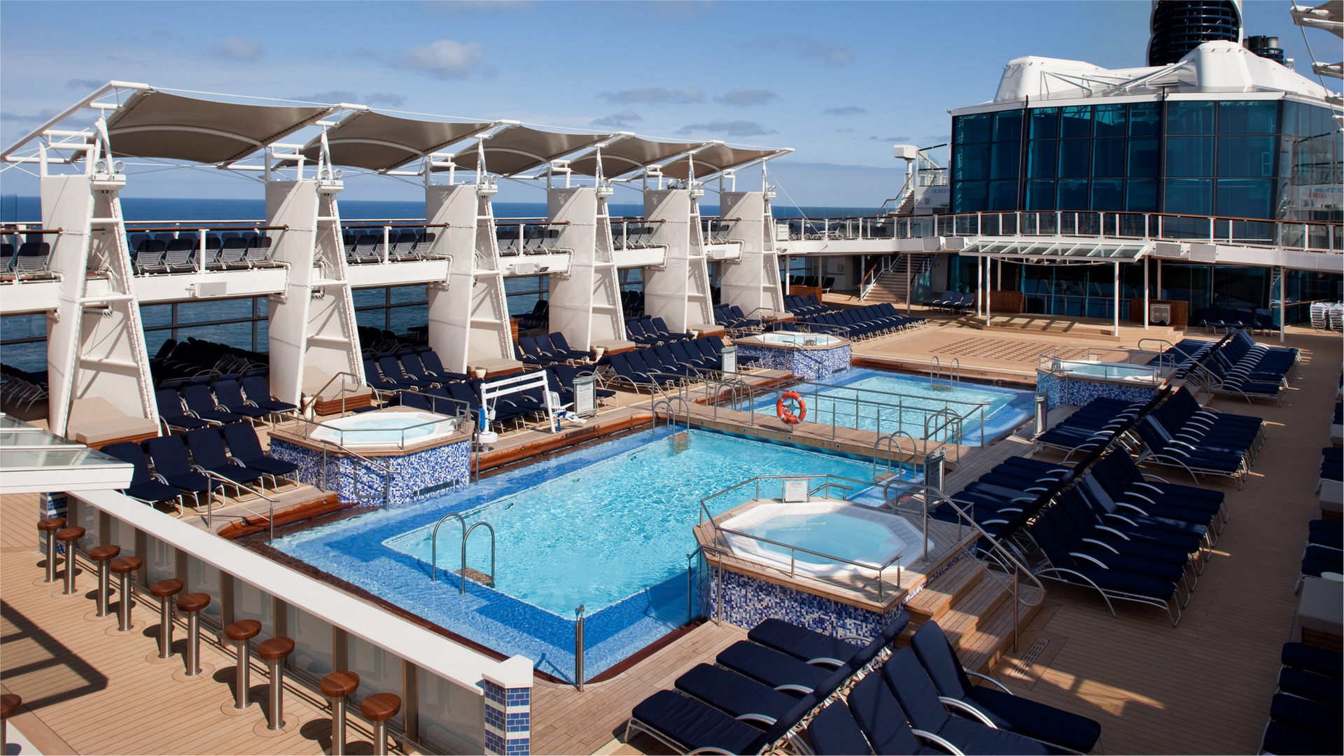 Cruise ship deck with pools, hot tubs, and rows of blue lounge chairs under canopies, on a sunny day.