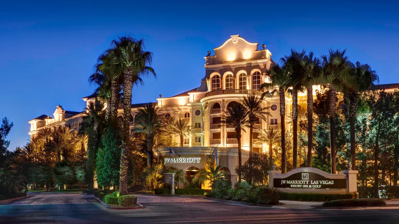 Hotel exterior at dusk, with a lighted sign and palm trees lining the driveway. Blue sky backdrop.
