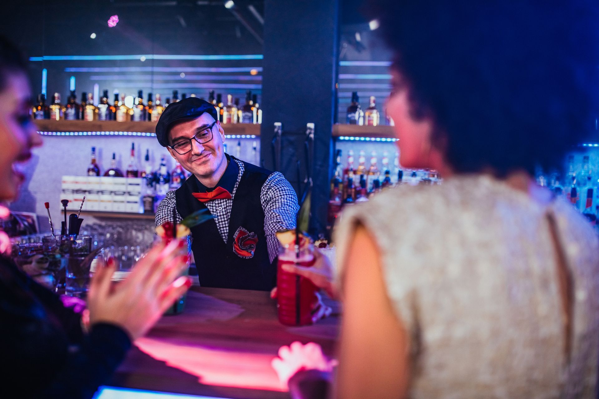 A bartender smiles, handing cocktails to two women at a bar with colorful lights and liquor bottles.