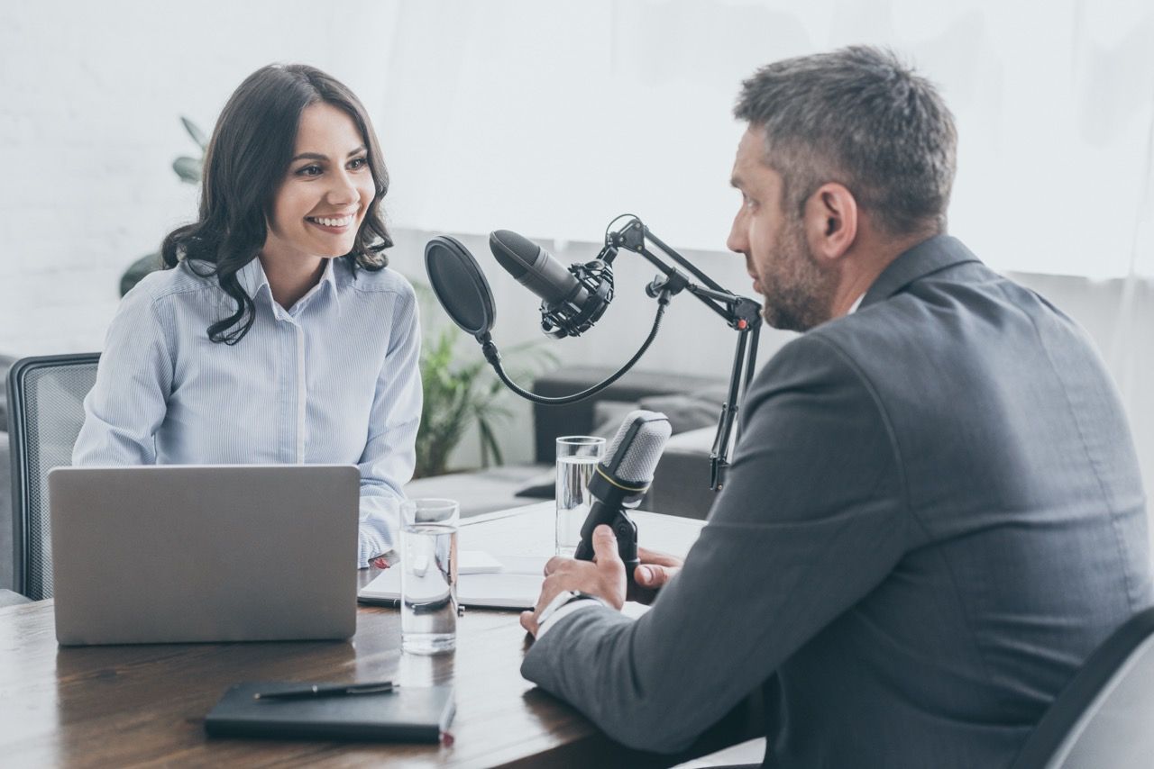 Woman and man in a podcast studio; the woman smiles at the man. They sit across from each other at a table with microphones and a laptop.