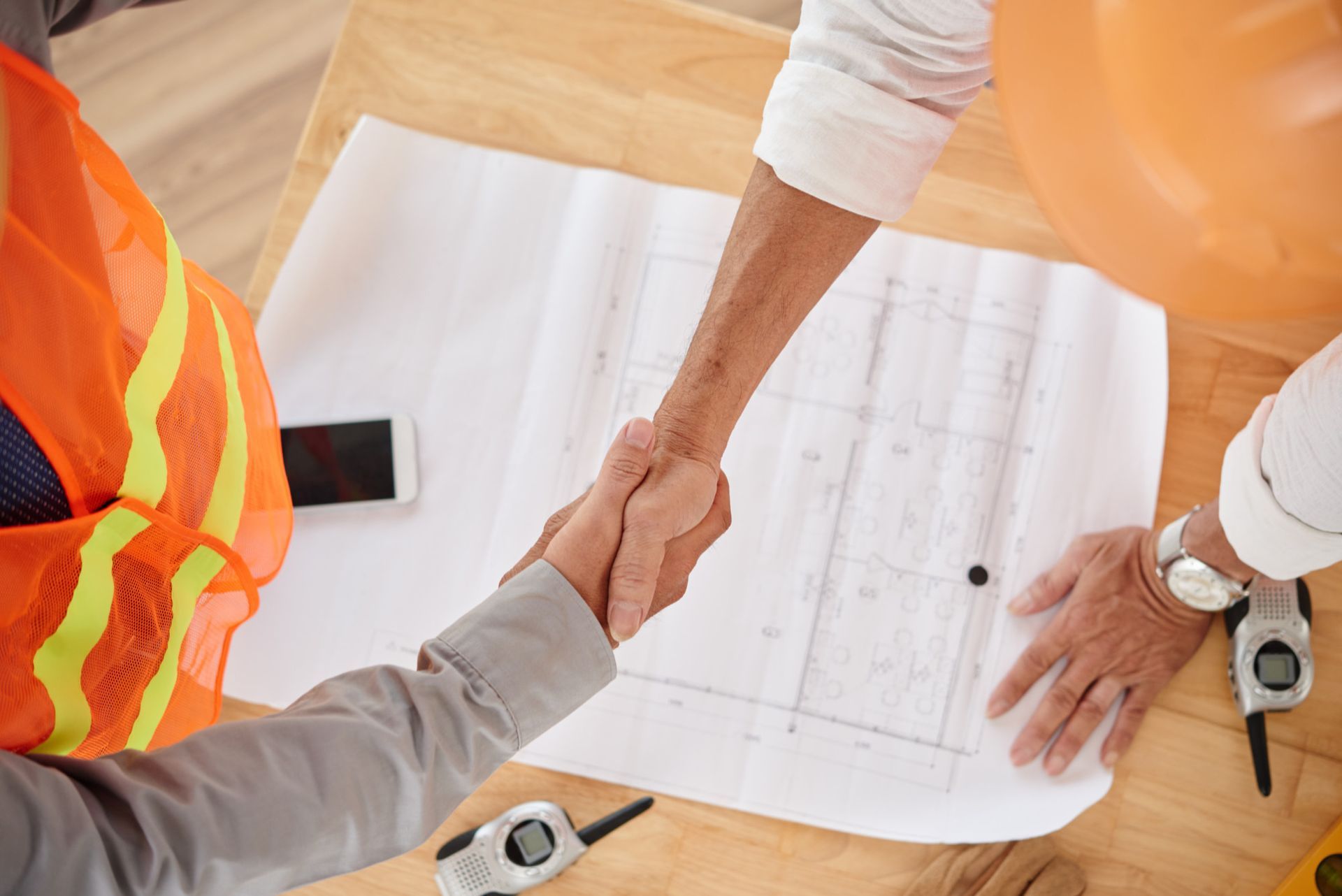 Construction workers shaking hands over a blueprint, wearing safety vests and hard hats.