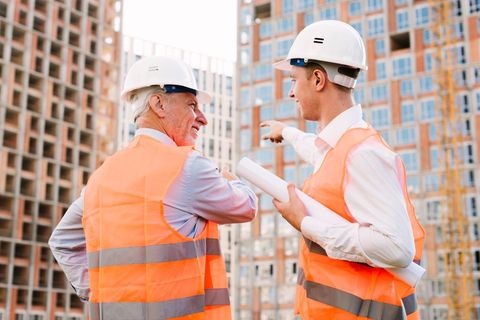 Two construction workers in safety vests and hard hats at a building site, one pointing, discussing plans.