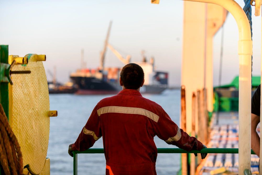 Person in red uniform looking at cargo ships near a port, from the deck of another vessel.
