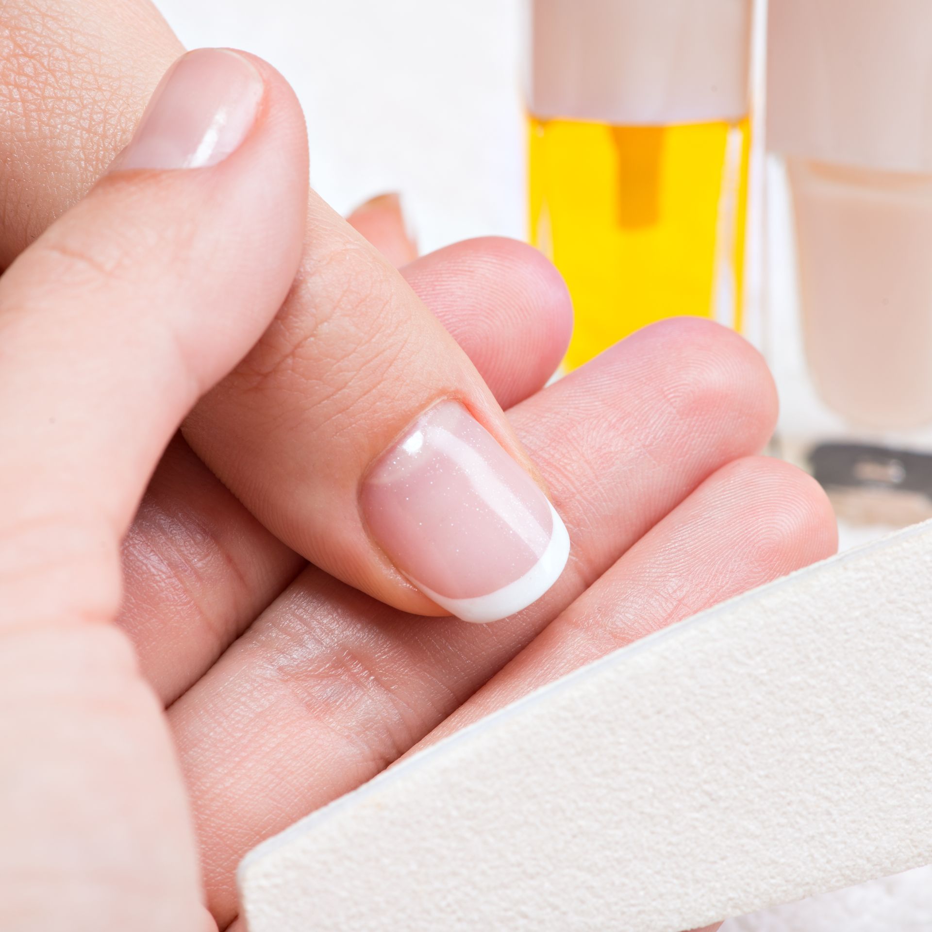 Nail being filed with a white emery board, French manicure on a hand, bottles of nail products in background.