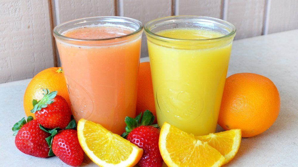 Two glasses of juice surrounded by oranges and strawberries on a table.