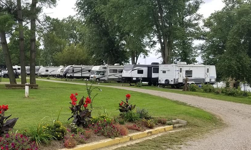 A gravel driveway lined with colorful flowers leads to a row of RVs parked on a grassy lot with trees near a lake.
