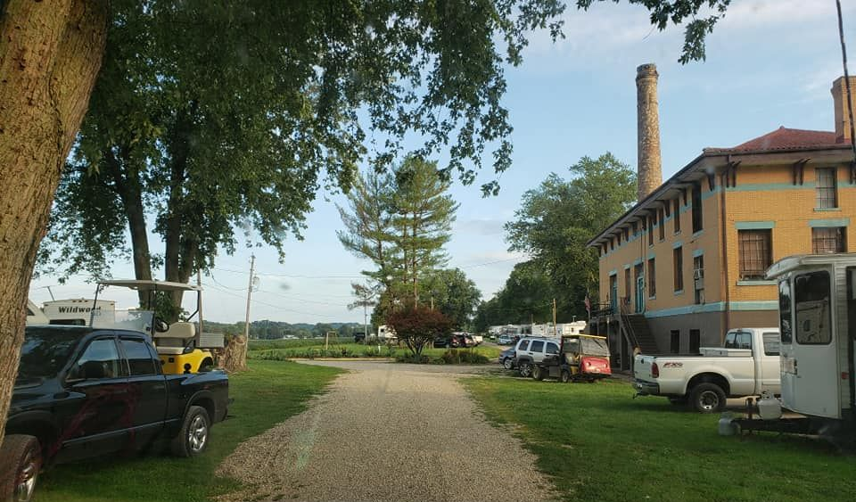 A gravel driveway leads toward trees and a yellow building with a tall brick chimney, flanked by parked trucks and trailers.