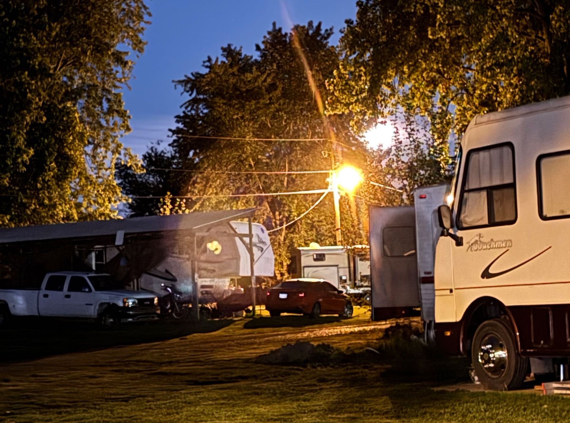 RVs and cars parked at a campsite at dusk, with warm lights glowing under trees