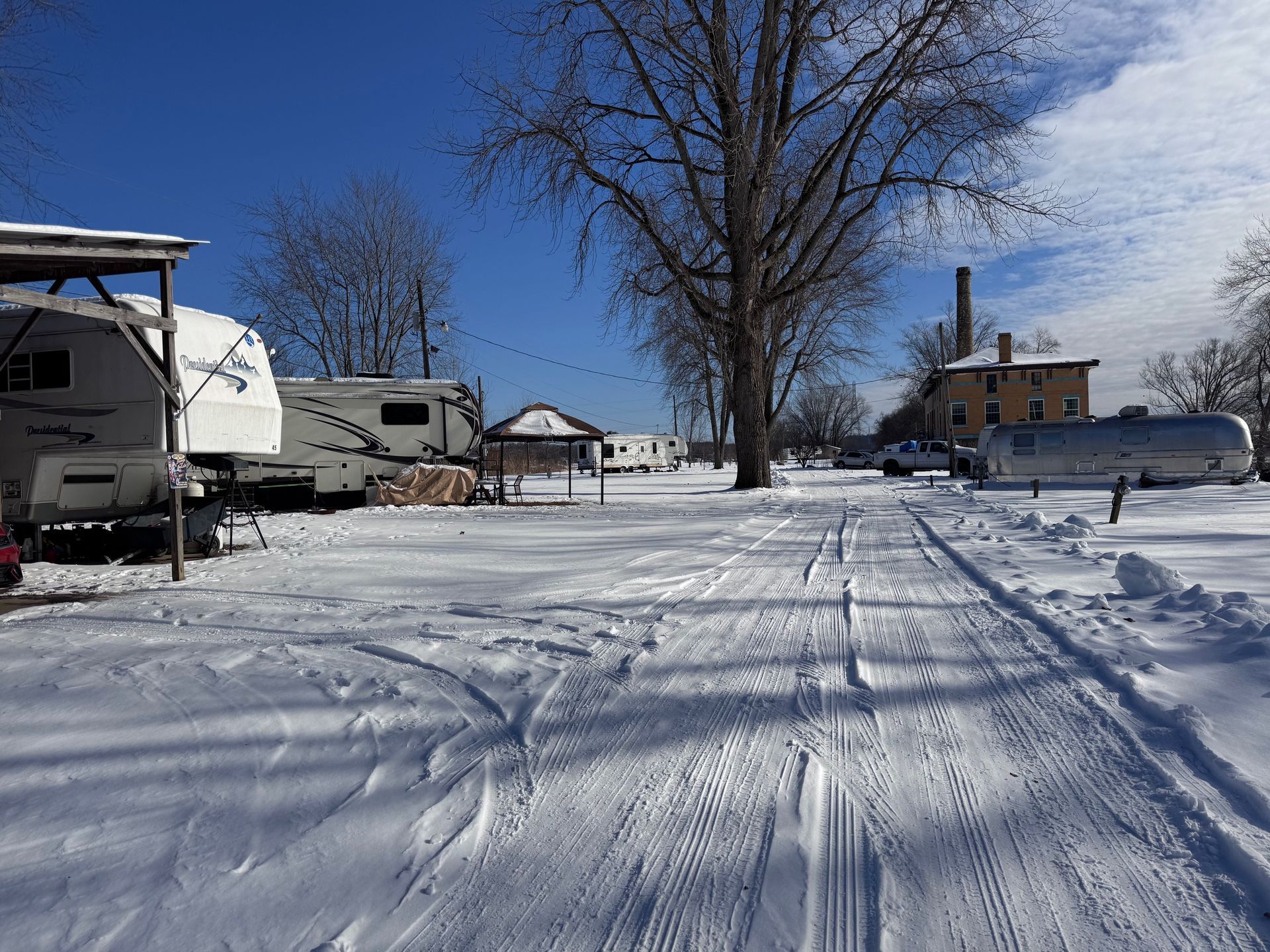 Snow-covered rural driveway with RVs, a bare tree, and a house under a clear blue sky.