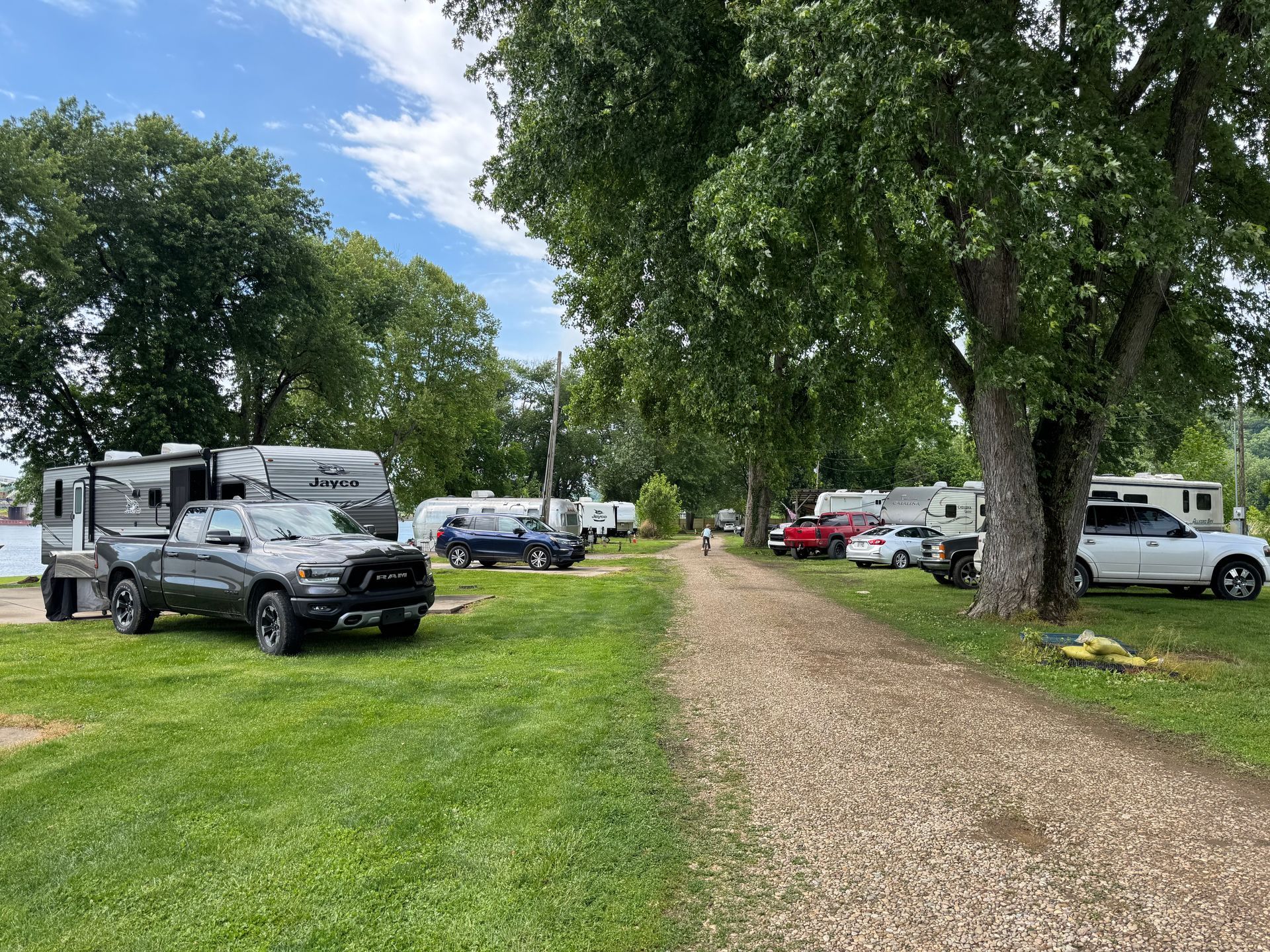 Gravel path through a tree-lined RV park with parked cars and trailers on a sunny day