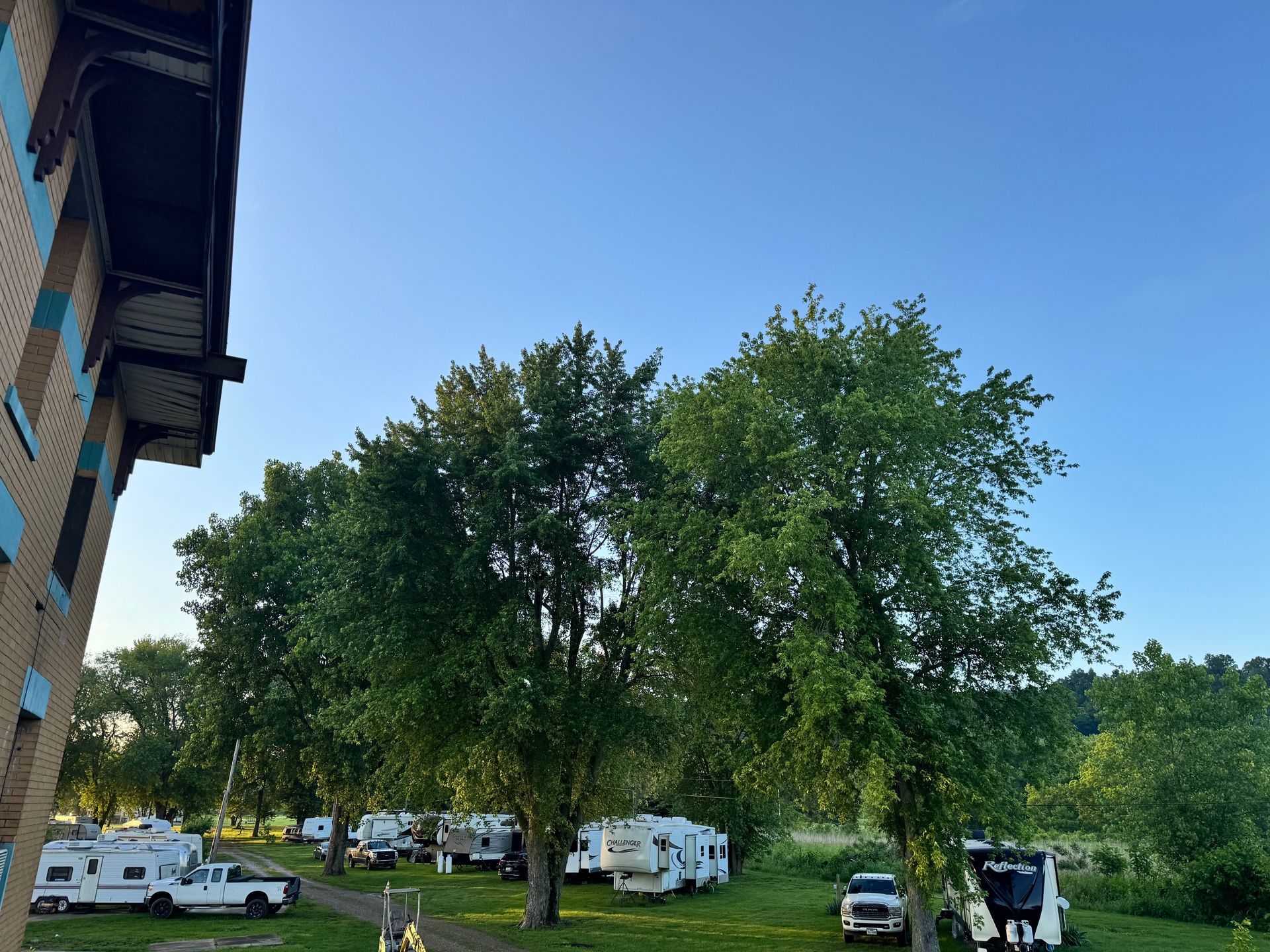 Tree-filled RV campsite with parked campers, a truck, and a building under a clear blue sky