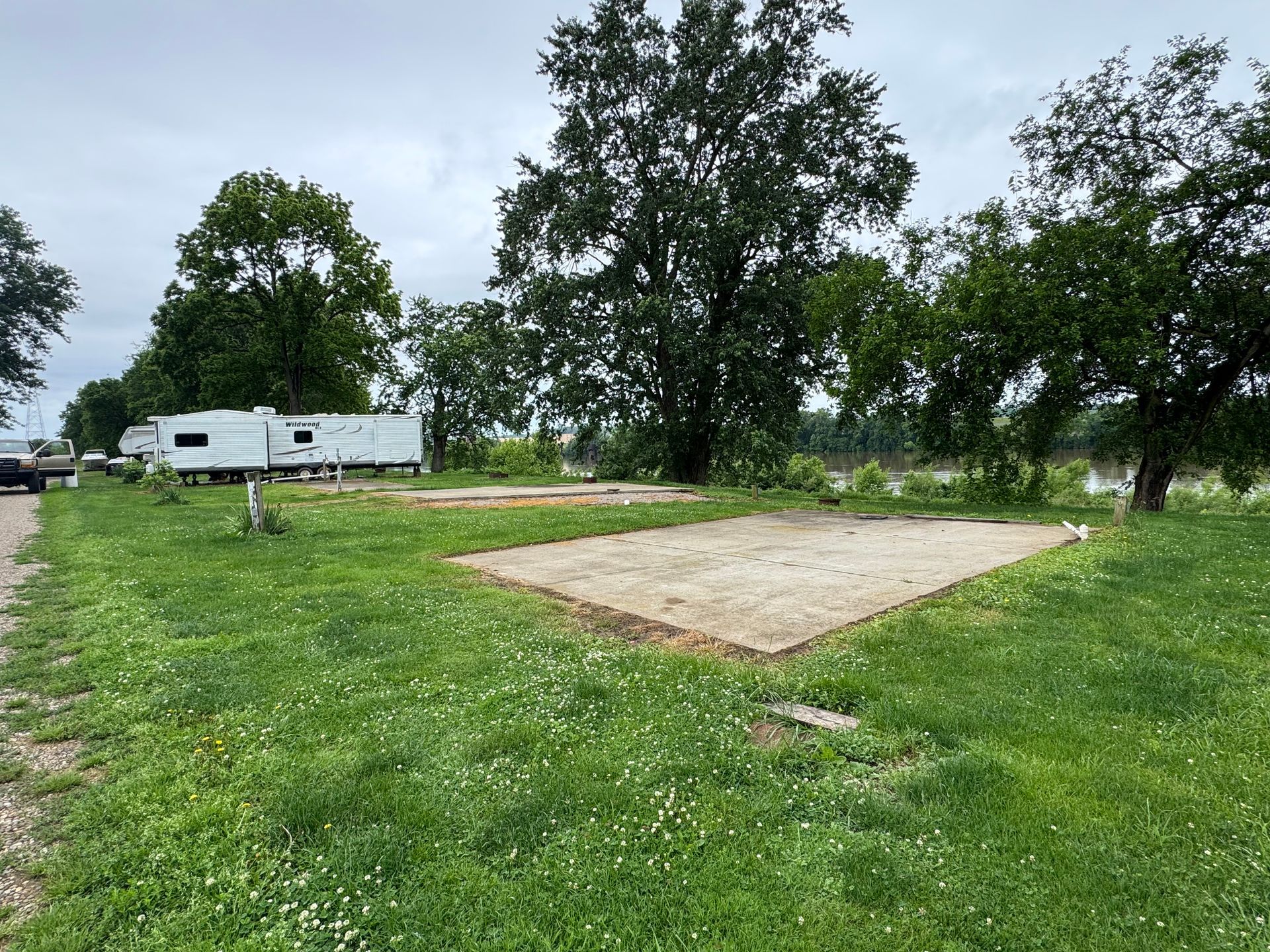 Grassy campsite with a concrete pad, trees, and a white RV trailer in the background