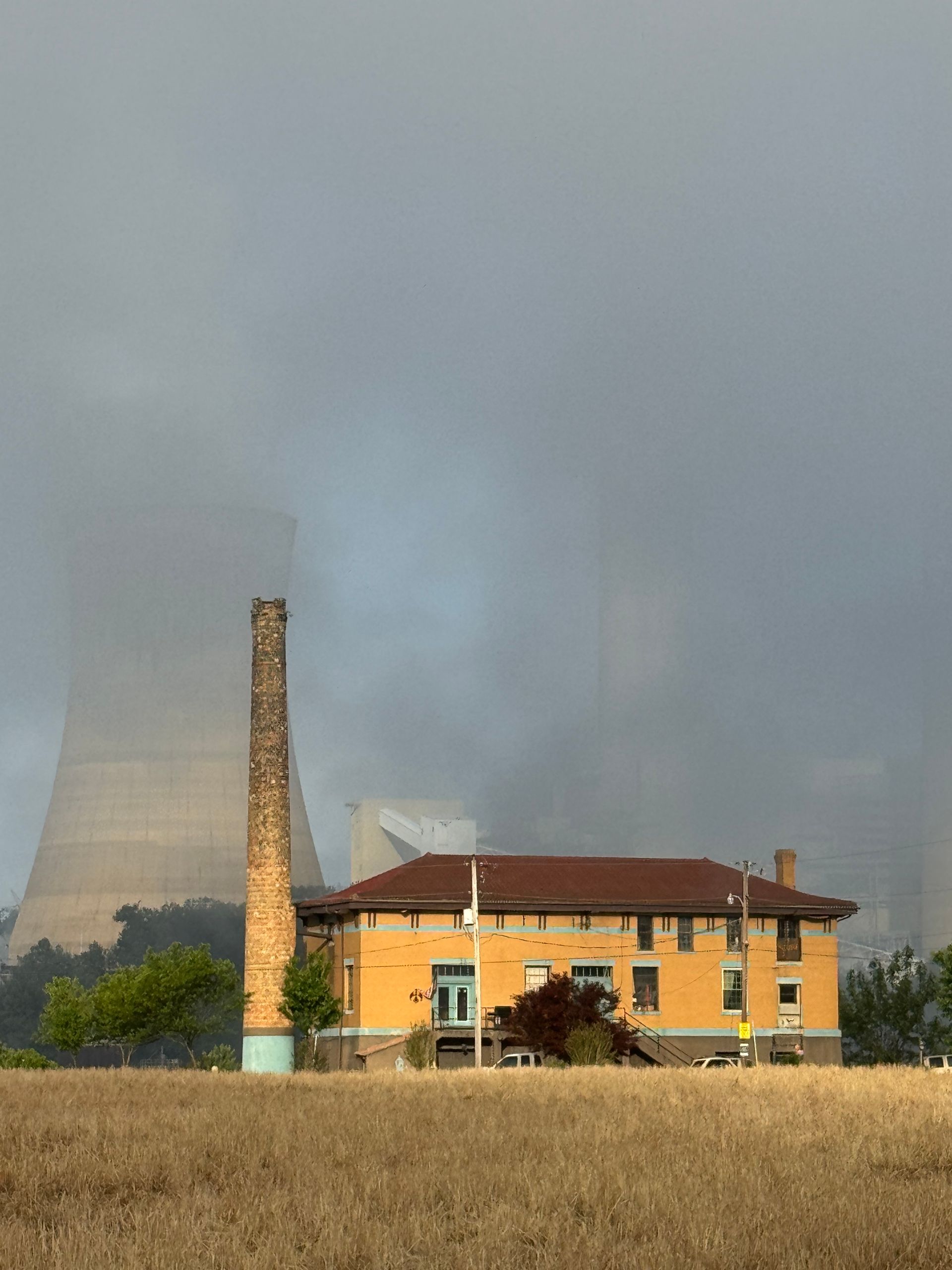 Yellow house and tall chimney in a dry field, with a power plant cooling tower in the foggy background