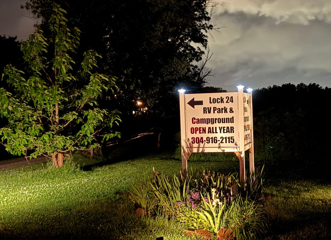 Lit trail sign at night near grassy path, with trees and flowers illuminated by a flashlight or car lights.