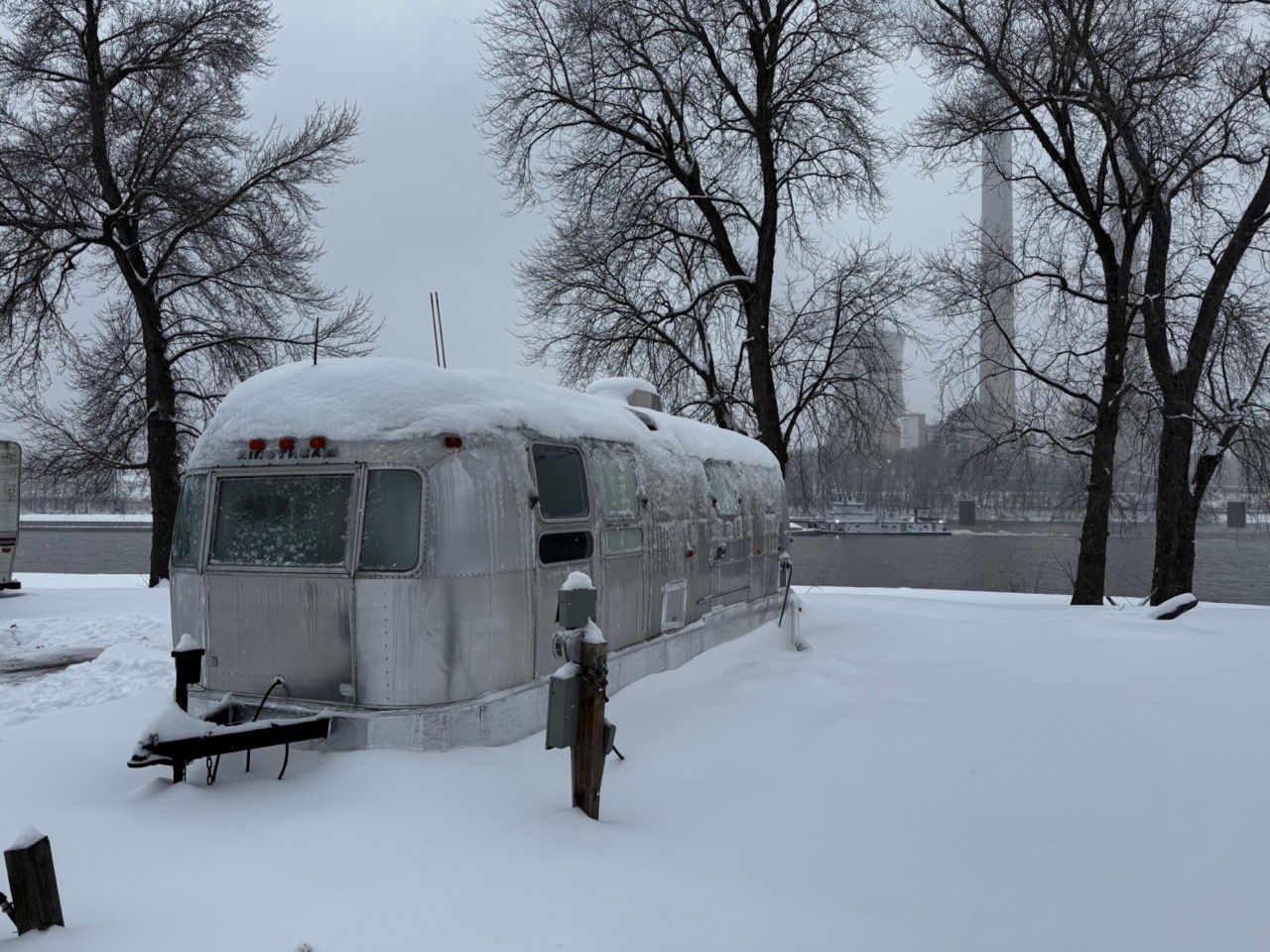 Snow-covered silver trailer in a winter park with bare trees and a city skyline in the distance