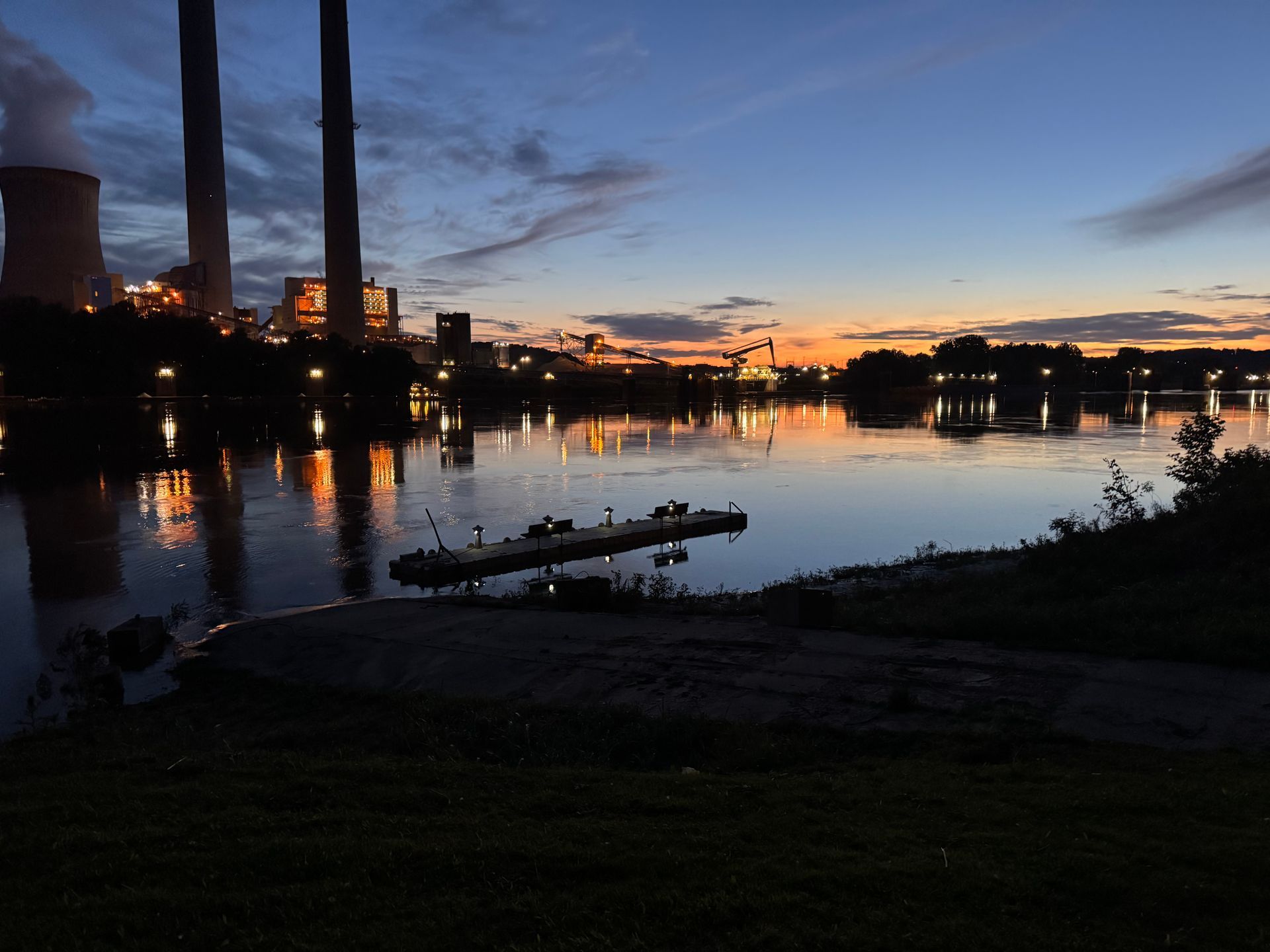 Sunset over a calm river with industrial smokestacks silhouetted on the left and glowing city lights in the distance