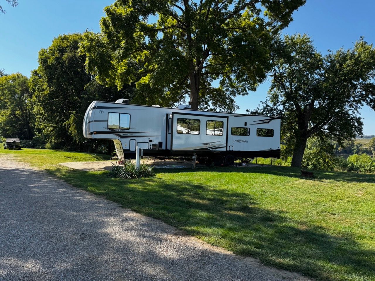 White travel trailer parked on a grassy campsite under large trees beside a gravel road