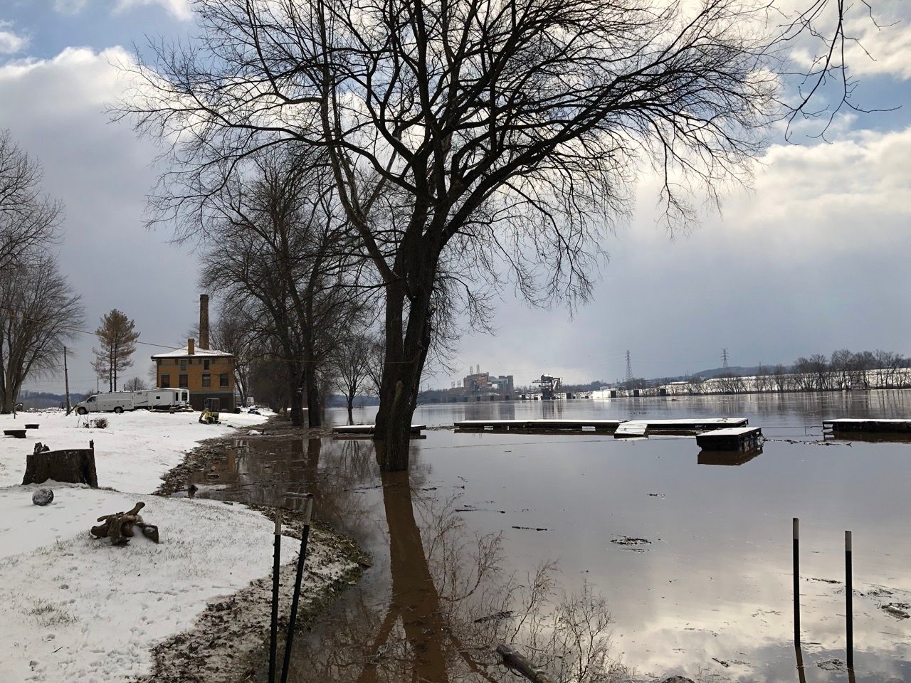 Snowy riverside with bare trees, calm water, and a small dock under a cloudy winter sky