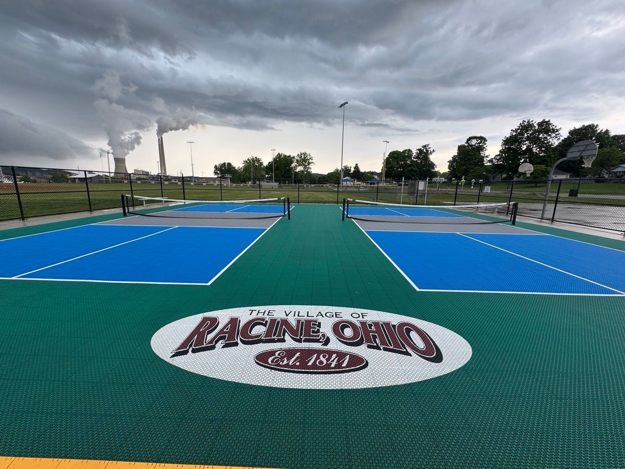 Outdoor blue pickleball courts with green center strip and a Racing Oval logo under a cloudy sky