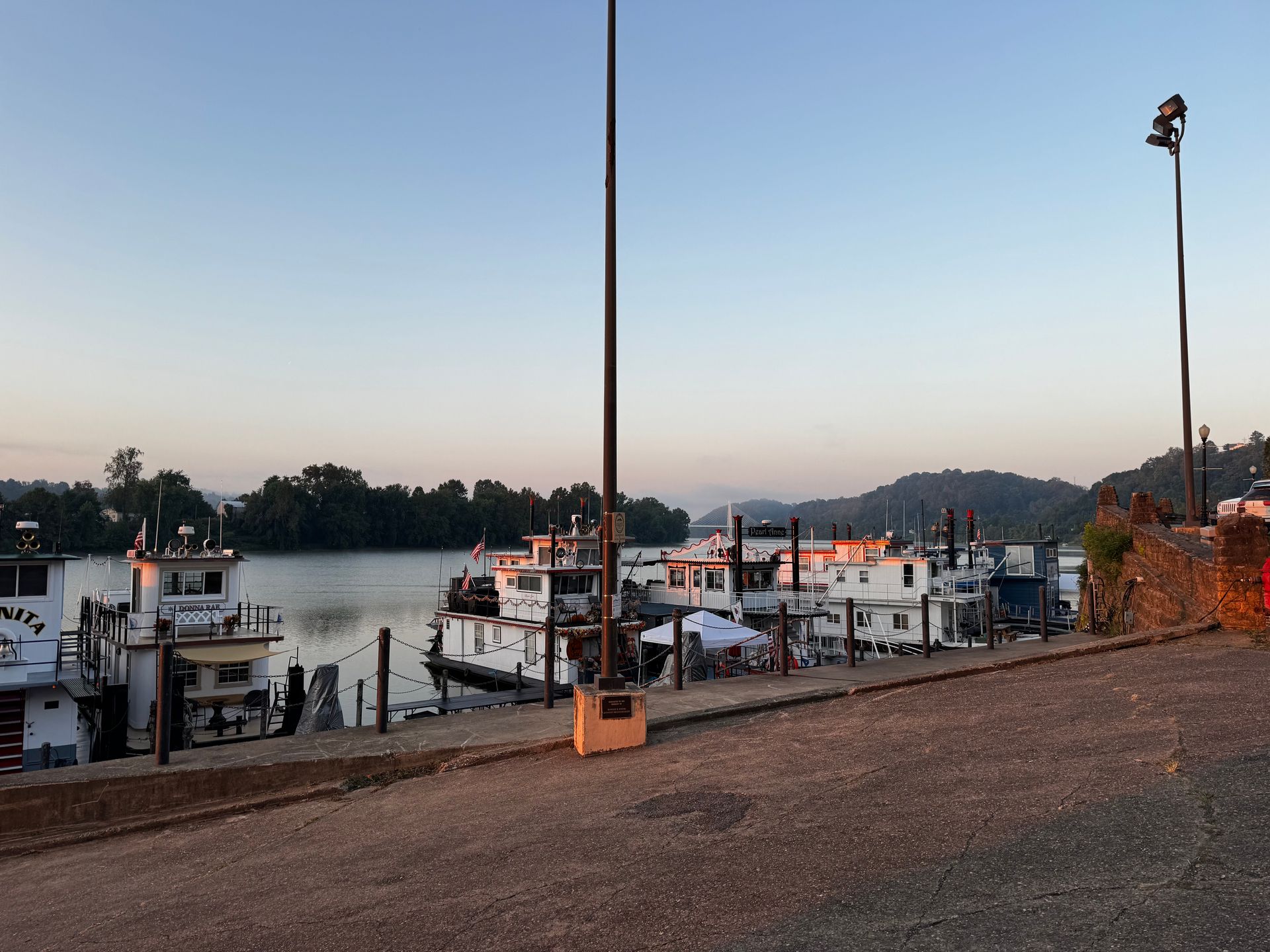 Dusk over a river with docked boats, a gravel bank, and tall lamp posts under a clear sky