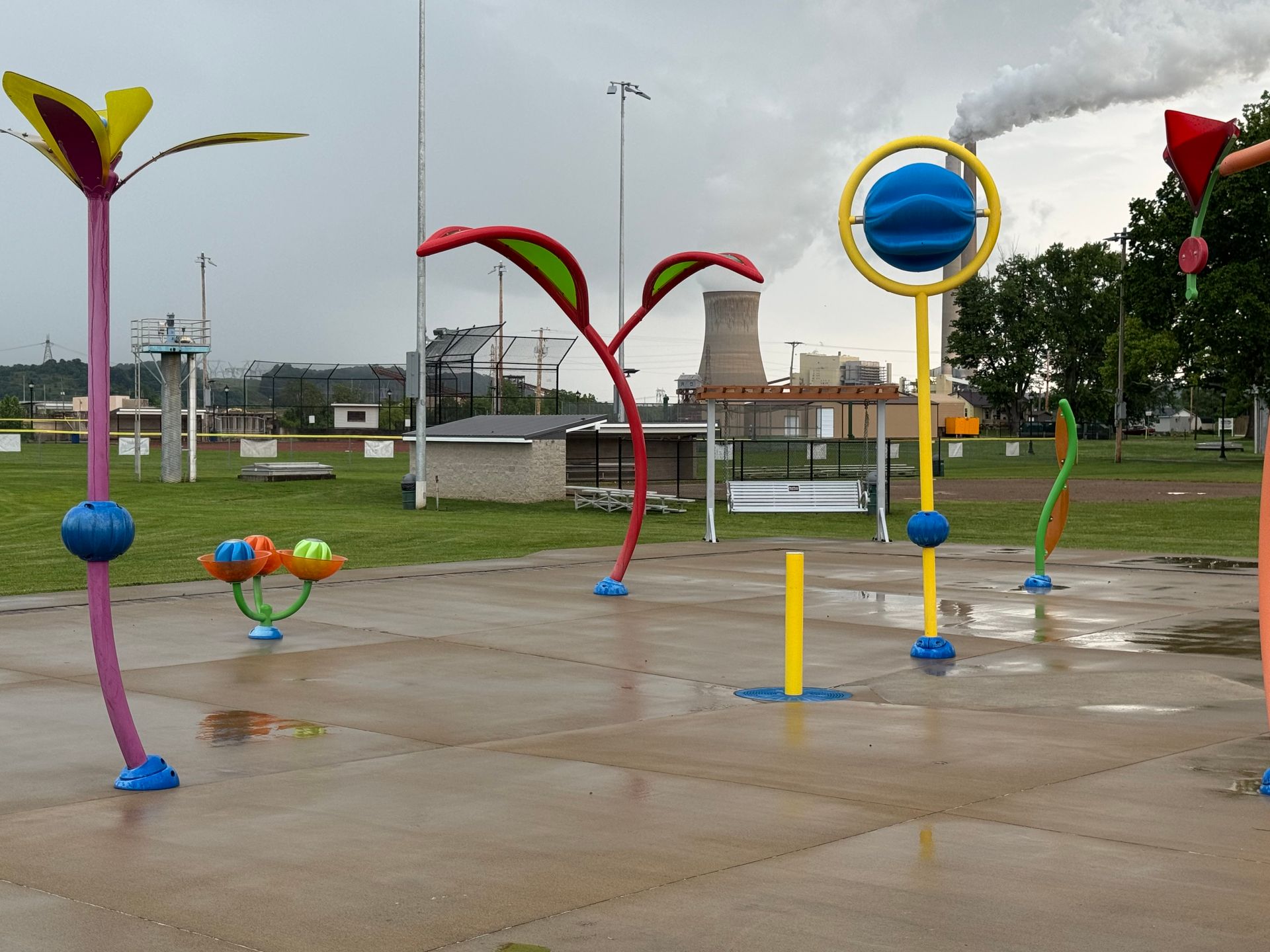 Colorful interactive water playground with sprinkler sculptures on a wet pavement under a cloudy sky