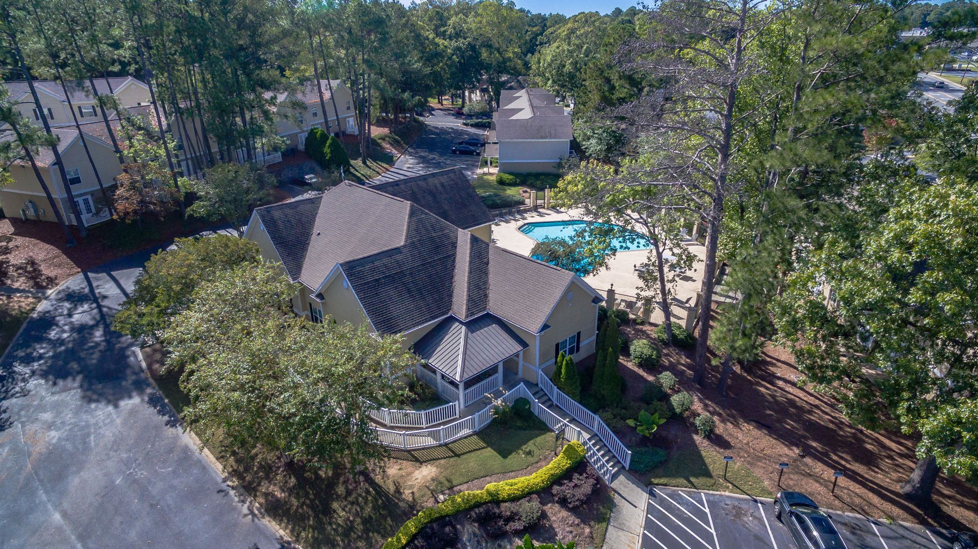 An aerial view of a tan clubhouse with a dark roof, adjacent to an outdoor swimming pool, surrounded by trees and paths.