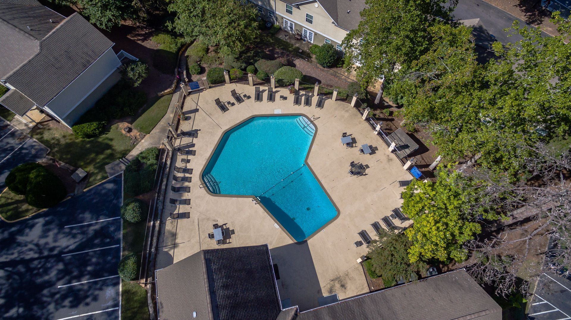 An aerial view of an outdoor swimming pool surrounded by a patio with lounge chairs and tables, set amidst trees.
