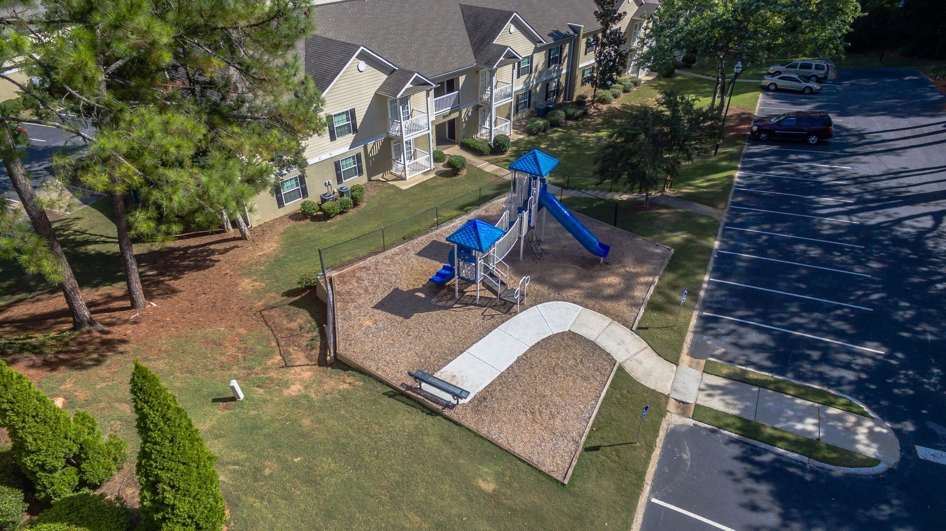 Aerial view of a small playground with blue equipment, mulch ground, and a white path near an apartment complex.