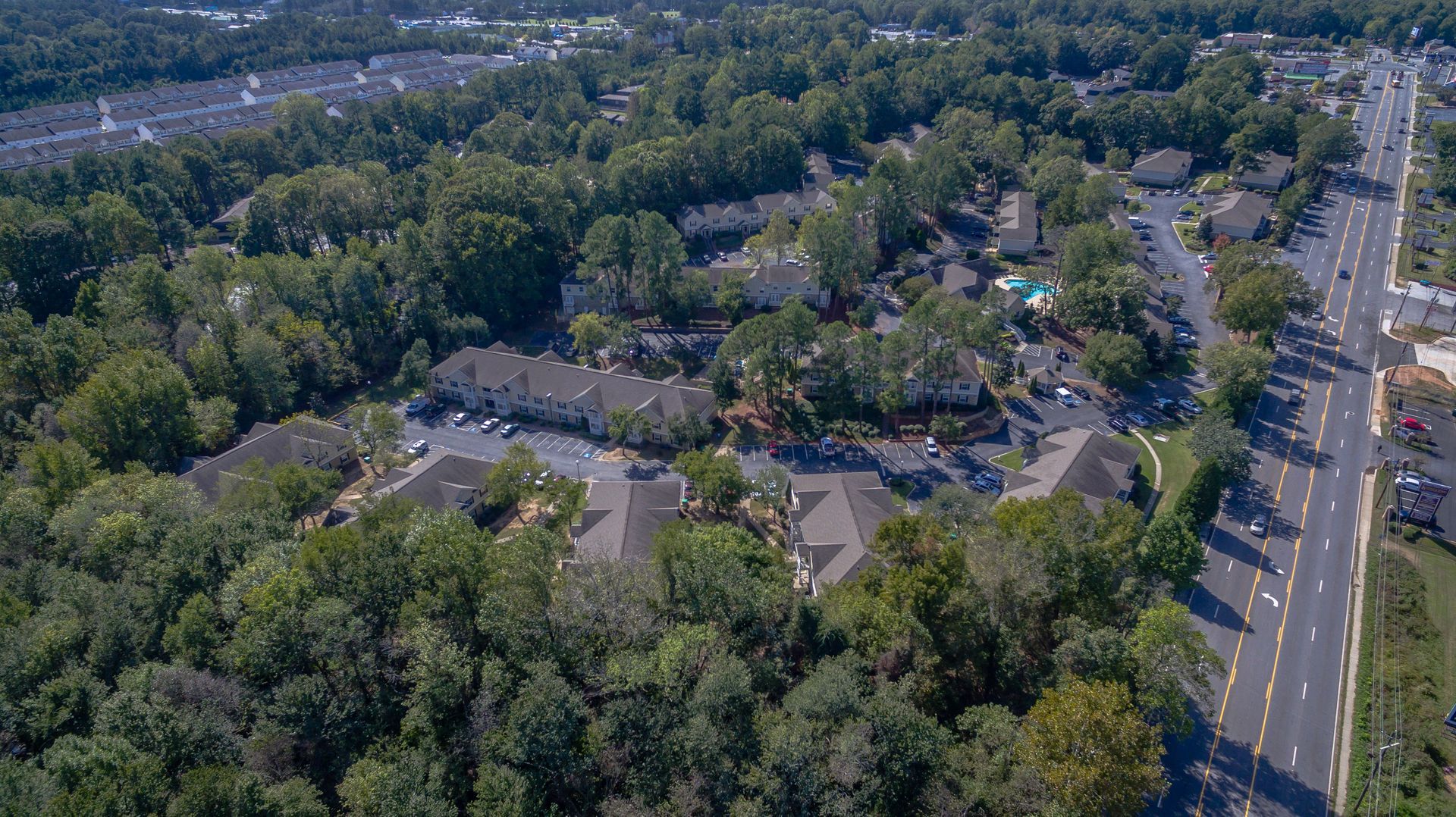 Aerial view of apartment complexes nestled among dense trees, adjacent to a multi-lane road in a suburban setting.
