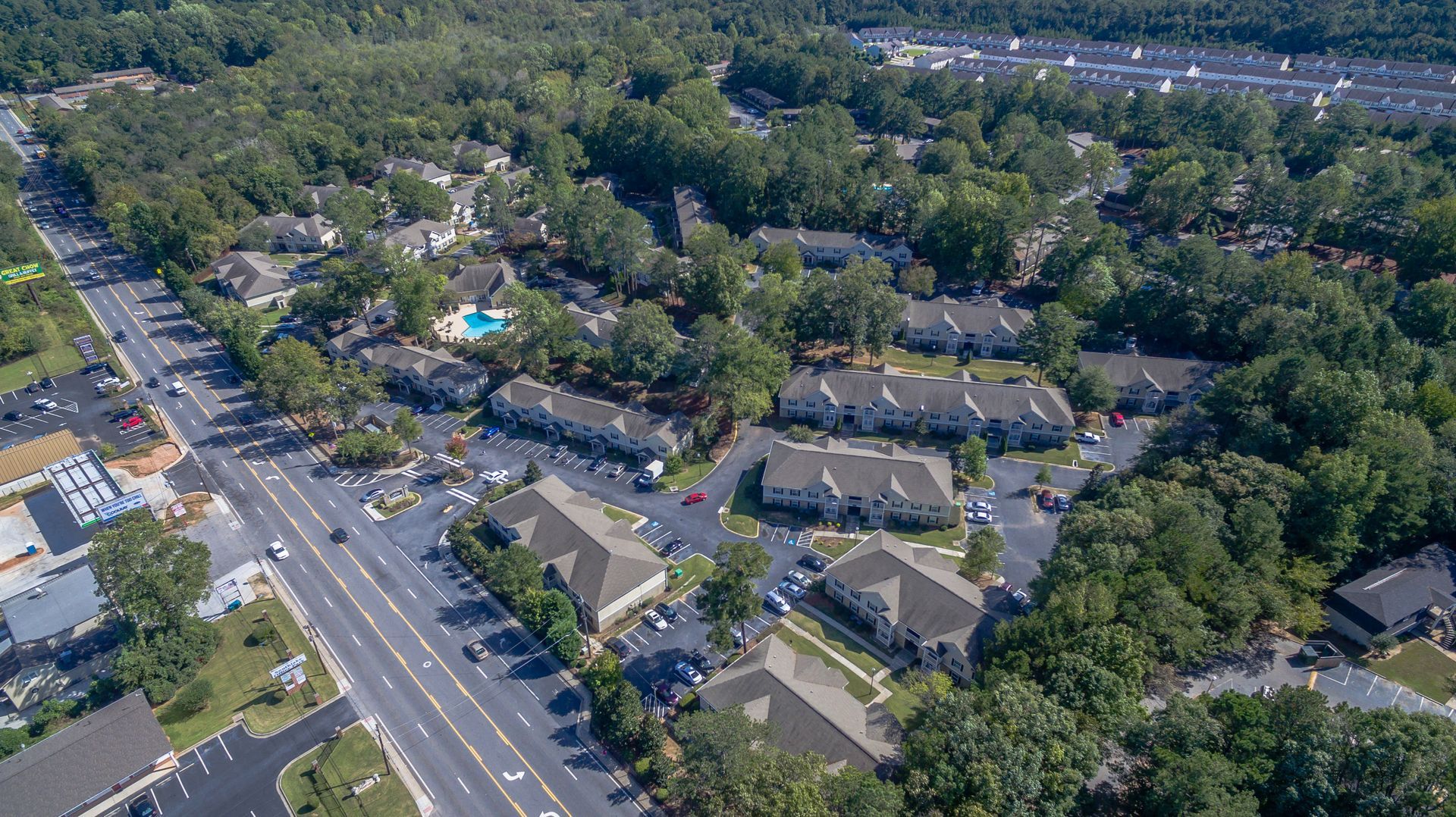 An aerial view of a multi-unit apartment complex nestled among dense trees, adjacent to a wide paved road.