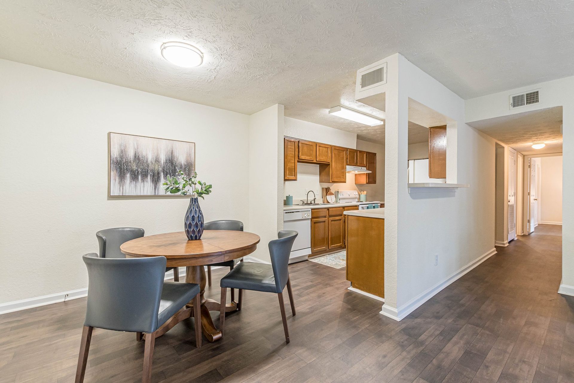 A dining area with a round wooden table, four gray chairs, and wall art, next to a galley kitchen with wooden cabinets.