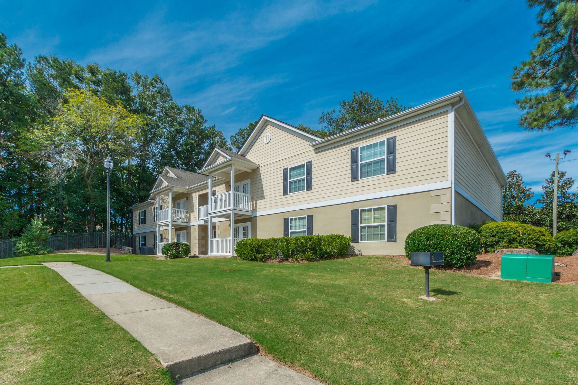 A multi-story beige apartment building exterior with white trim, surrounded by a lawn, trees, and a concrete path.