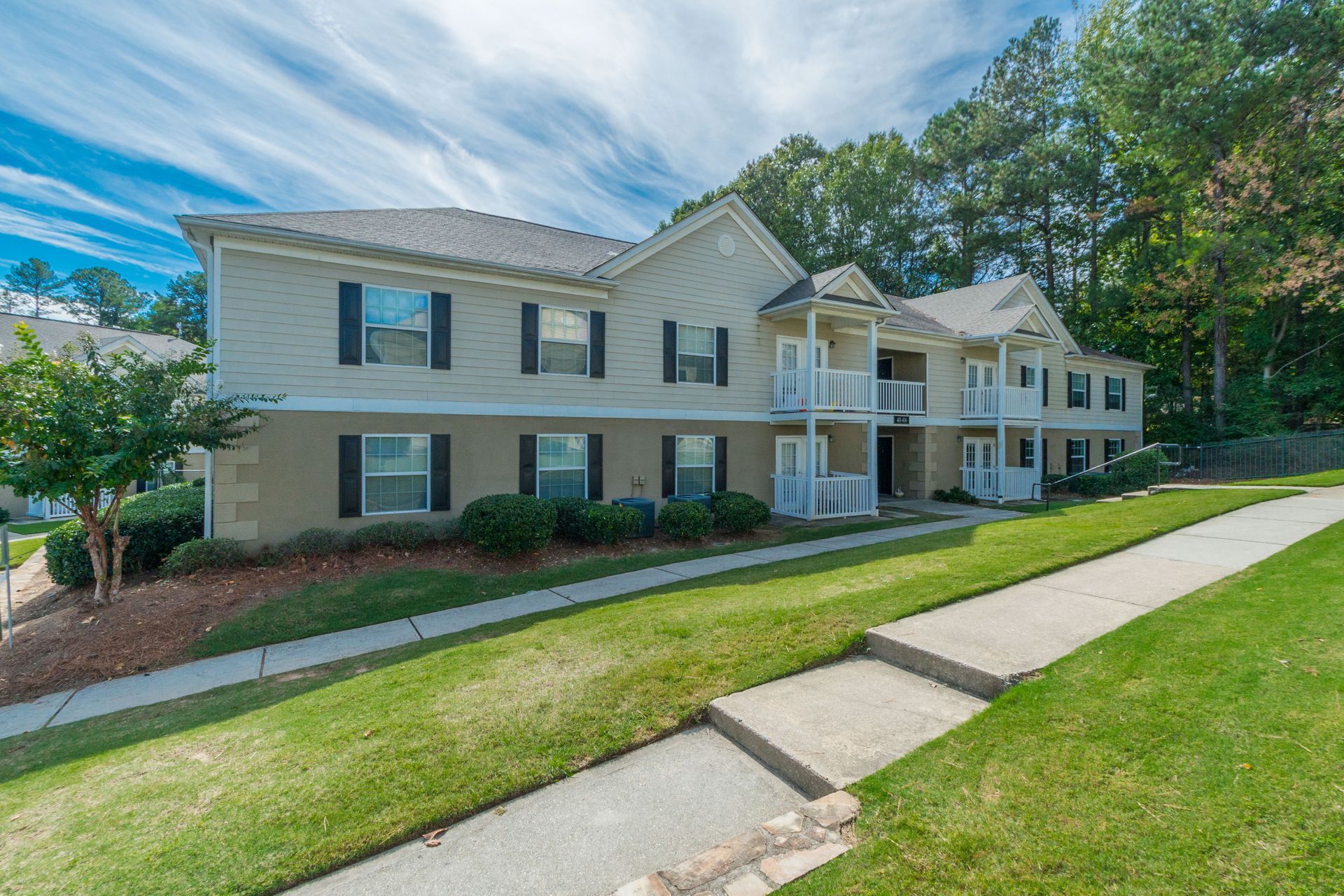 A two-story apartment complex with beige siding, dark shutters, white balconies, and green lawns under a blue sky.