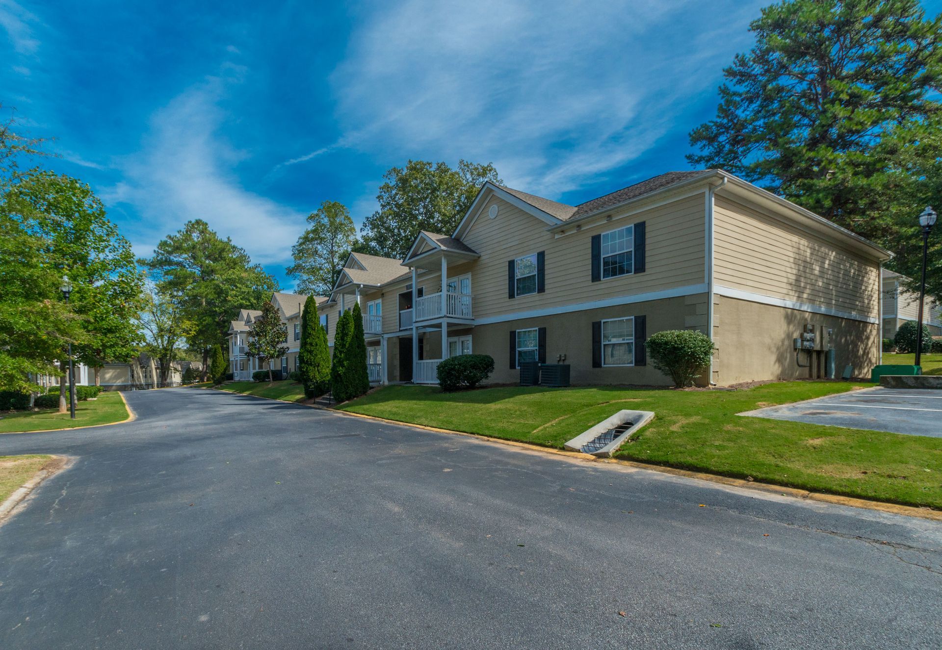 A tan two-story apartment building with dark shutters sits next to a paved road on a sunny day with blue skies.