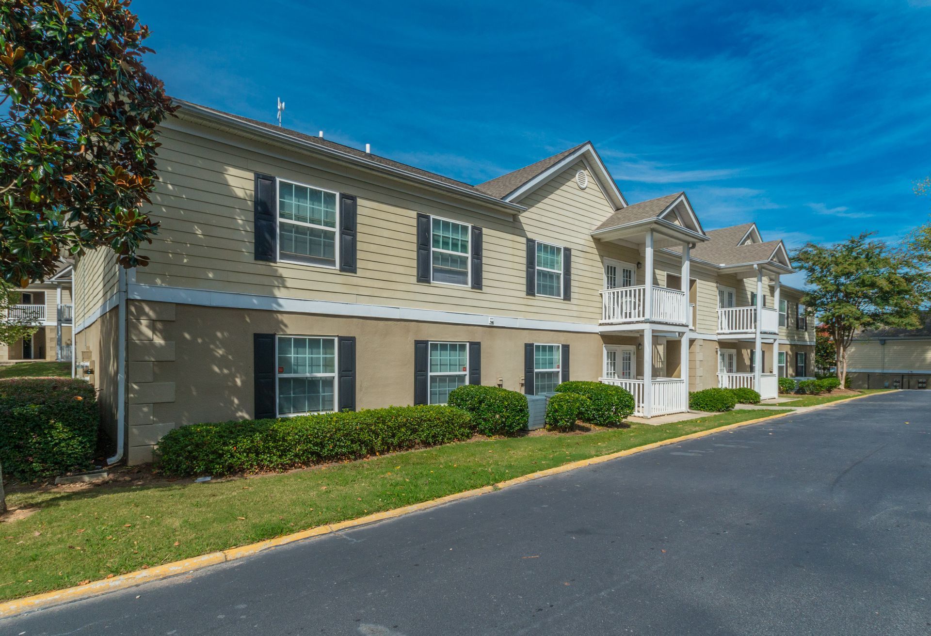 A two-story apartment building with beige siding, dark shutters, and white trim under a clear blue sky.