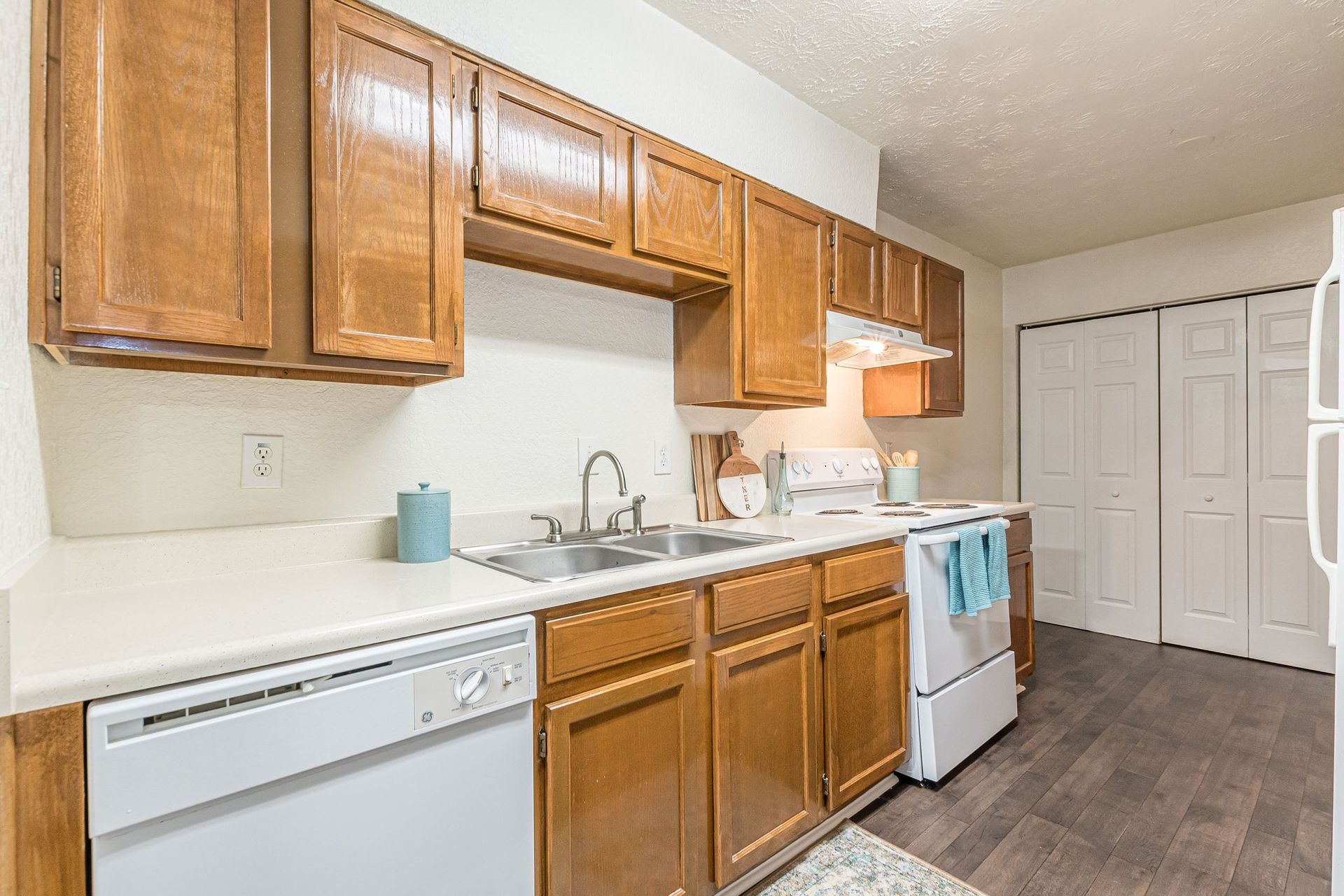 A galley-style kitchen featuring wood cabinets, white countertops, a white dishwasher, and a stove on wood-look flooring.