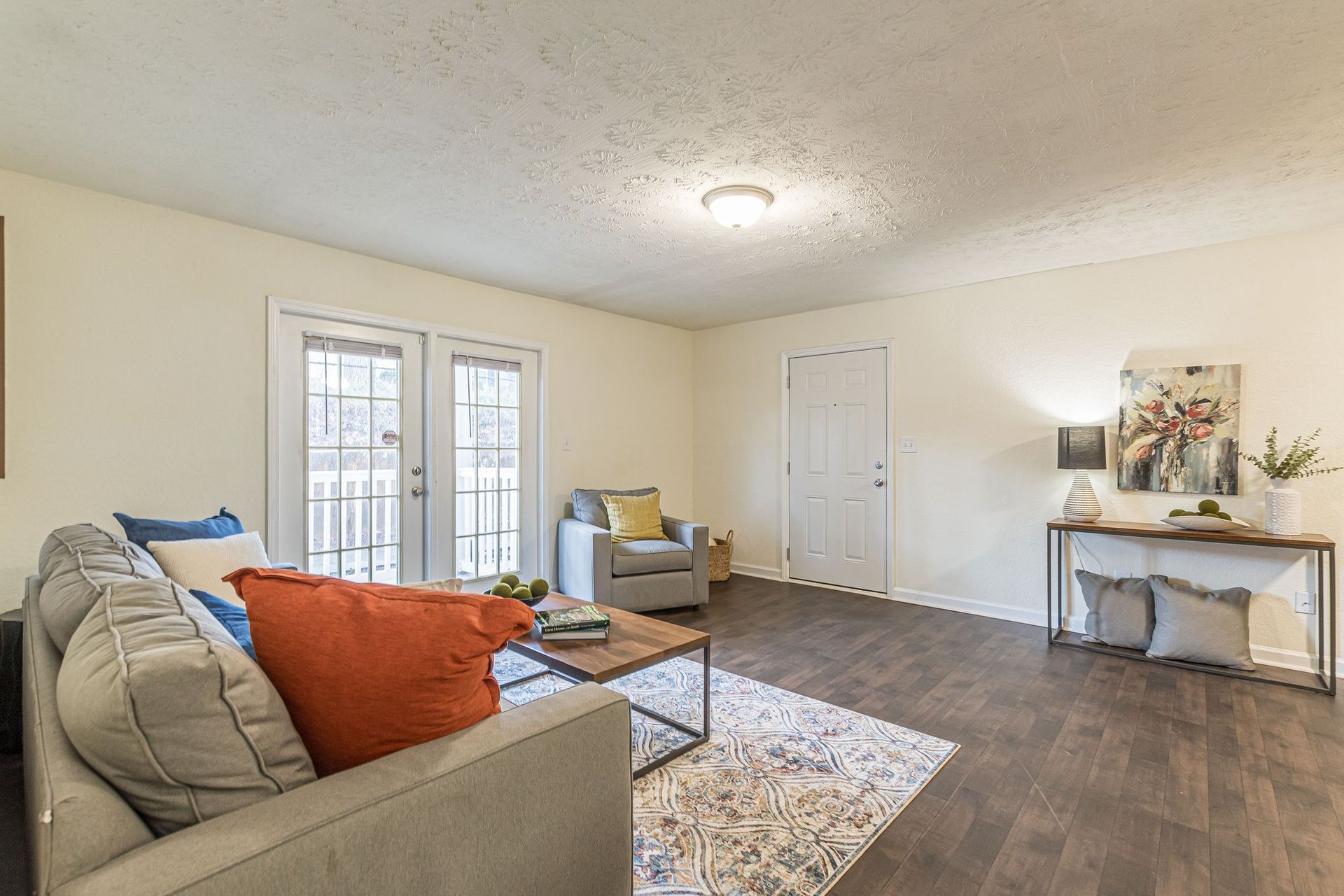 A furnished living room with a gray sofa, orange throw pillow, coffee table, area rug, and a console table by a white door.