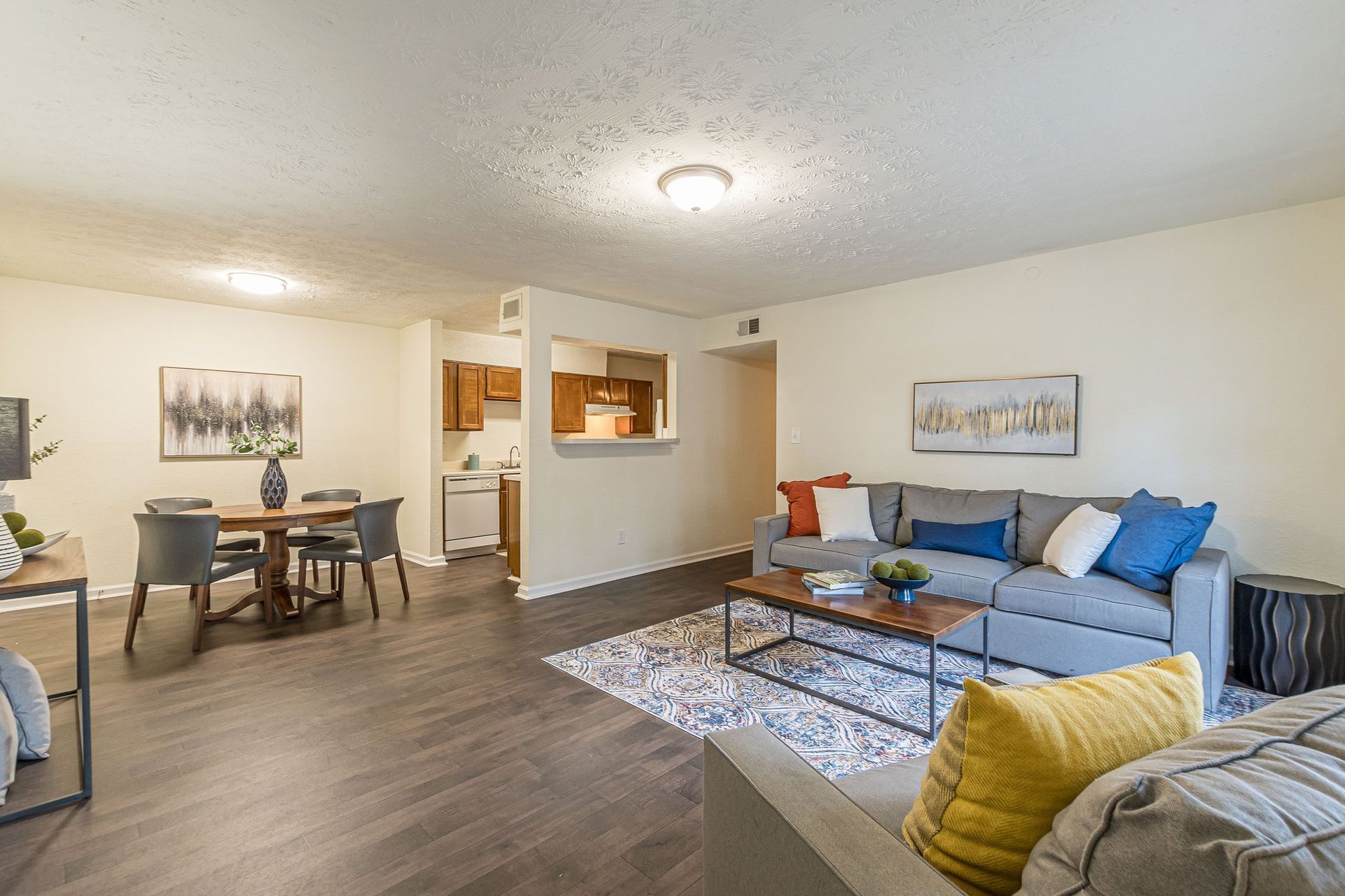 Living room with a gray sofa, coffee table, patterned rug, and dining area with a round table, open to a small kitchen.