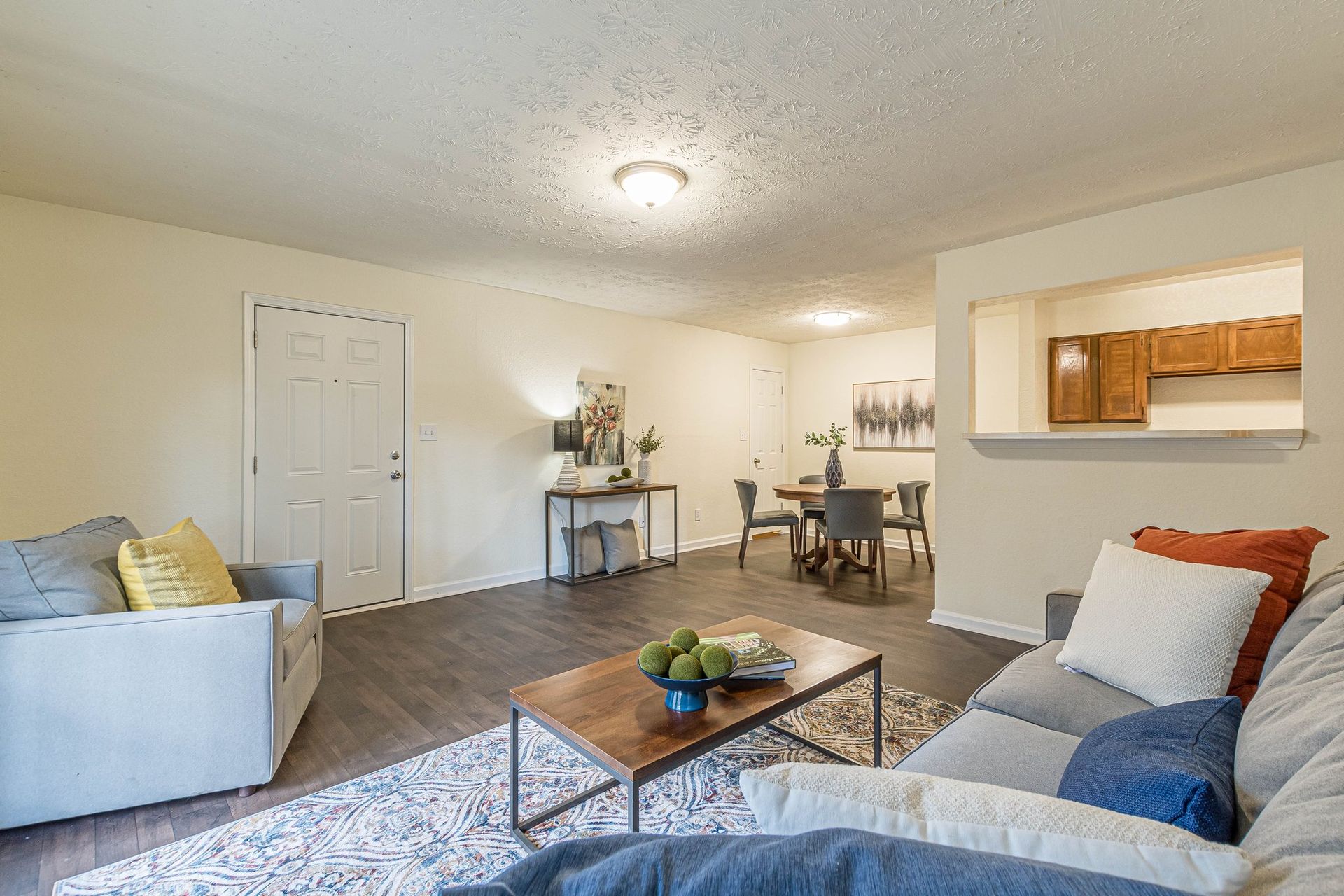 An open-concept living and dining room with a sofa, armchair, coffee table, and a kitchen pass-through window.