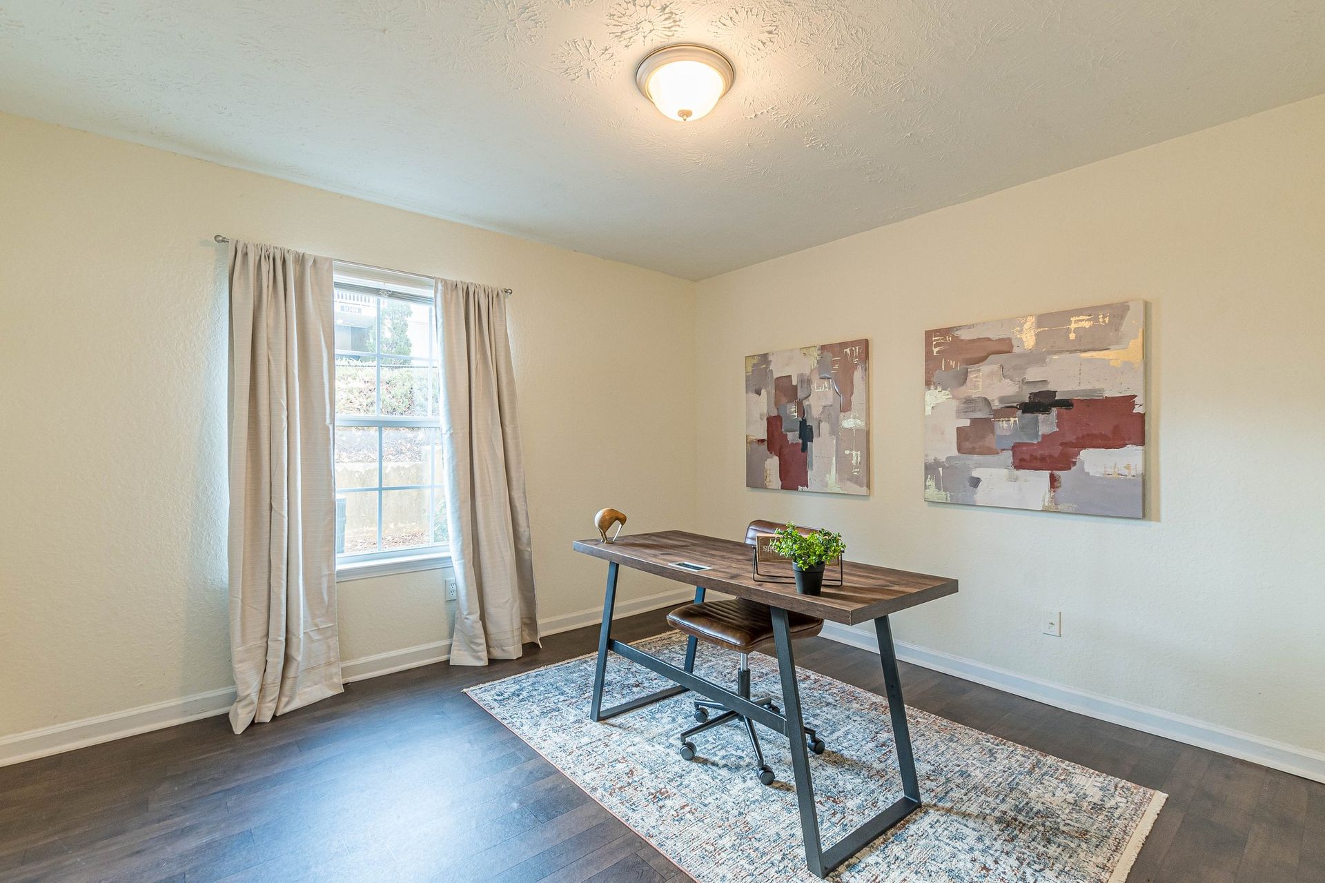 A home office with a wooden desk, office chair, and patterned rug, featuring white walls, curtains, and two wall paintings.