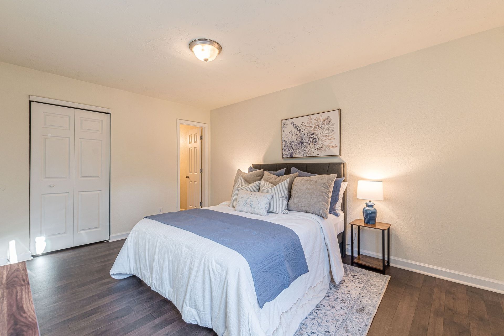 A brightly lit bedroom with a queen bed, blue and white bedding, dark wood flooring, and a small side table with a lamp.