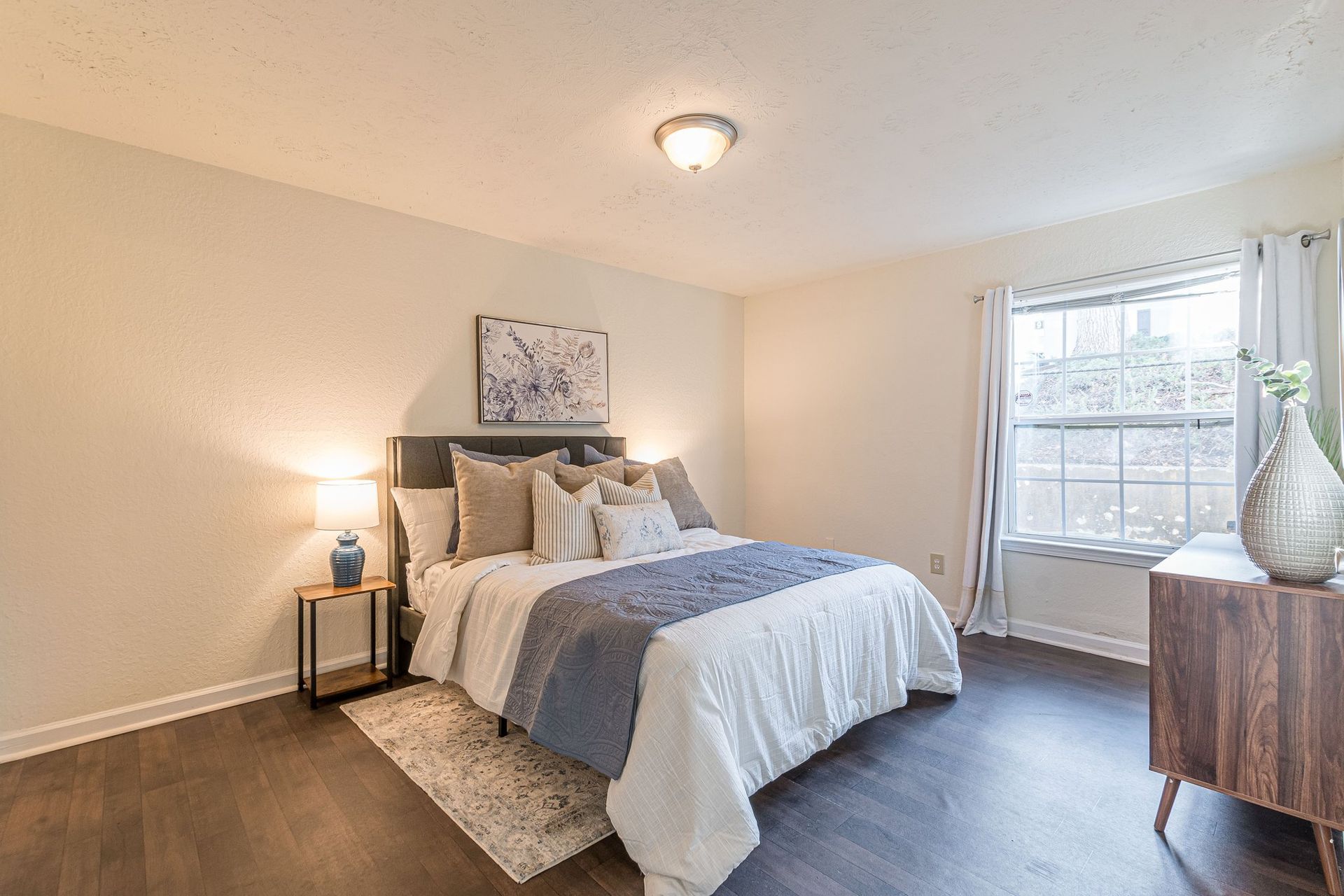 A bright bedroom featuring a queen bed with white linens, a blue throw blanket, two bedside tables, and a large window.