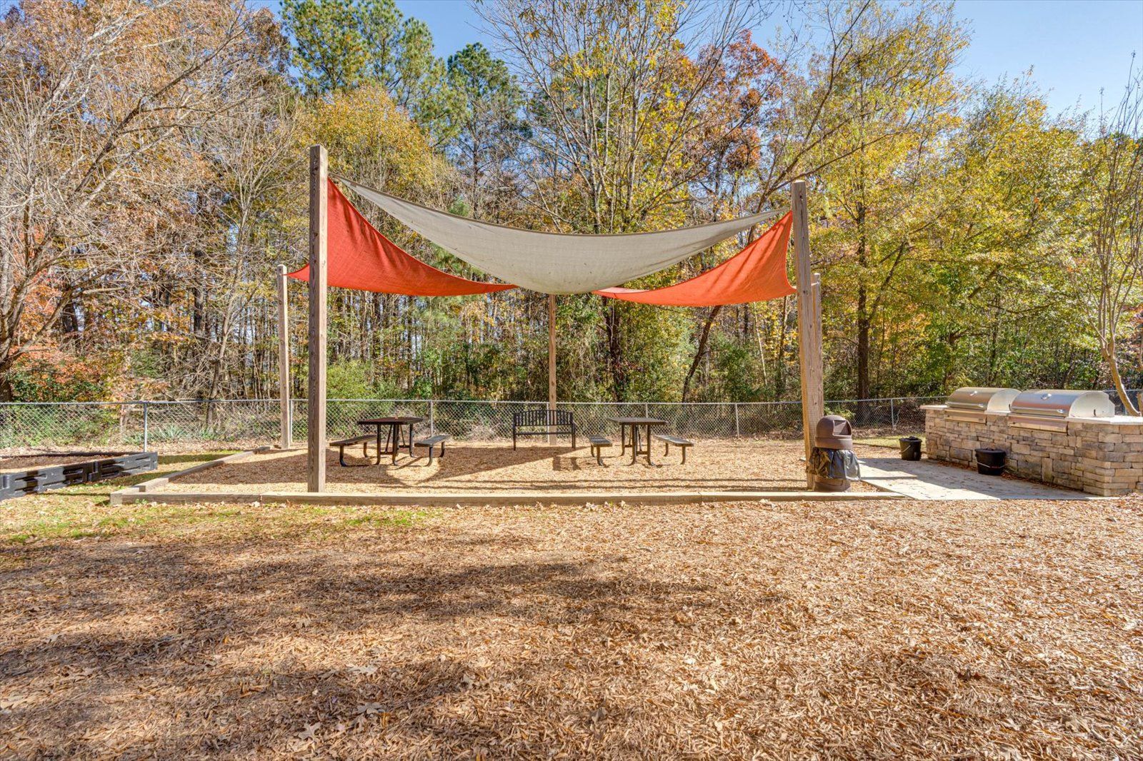 Outdoor pavilion with orange shade sails, picnic tables, and a stone fire pit in a wooded park