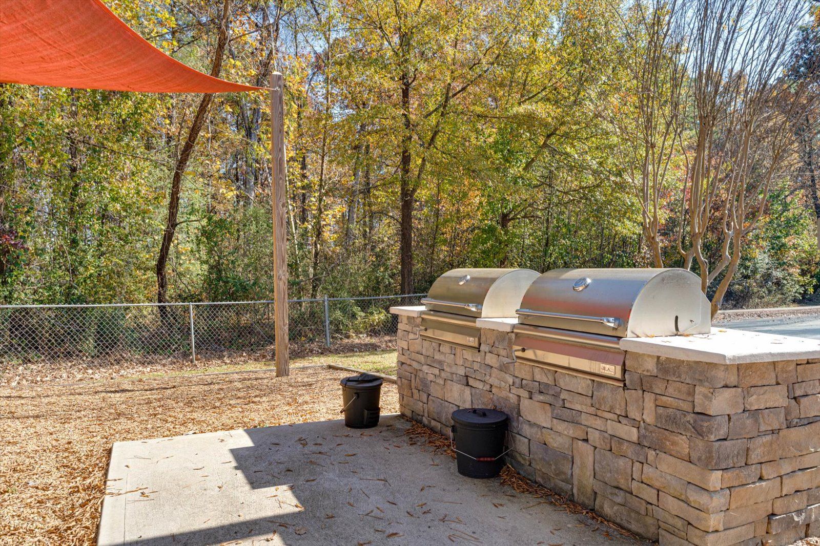 Outdoor stone grill area with stainless covers, black trash cans, and trees in the background