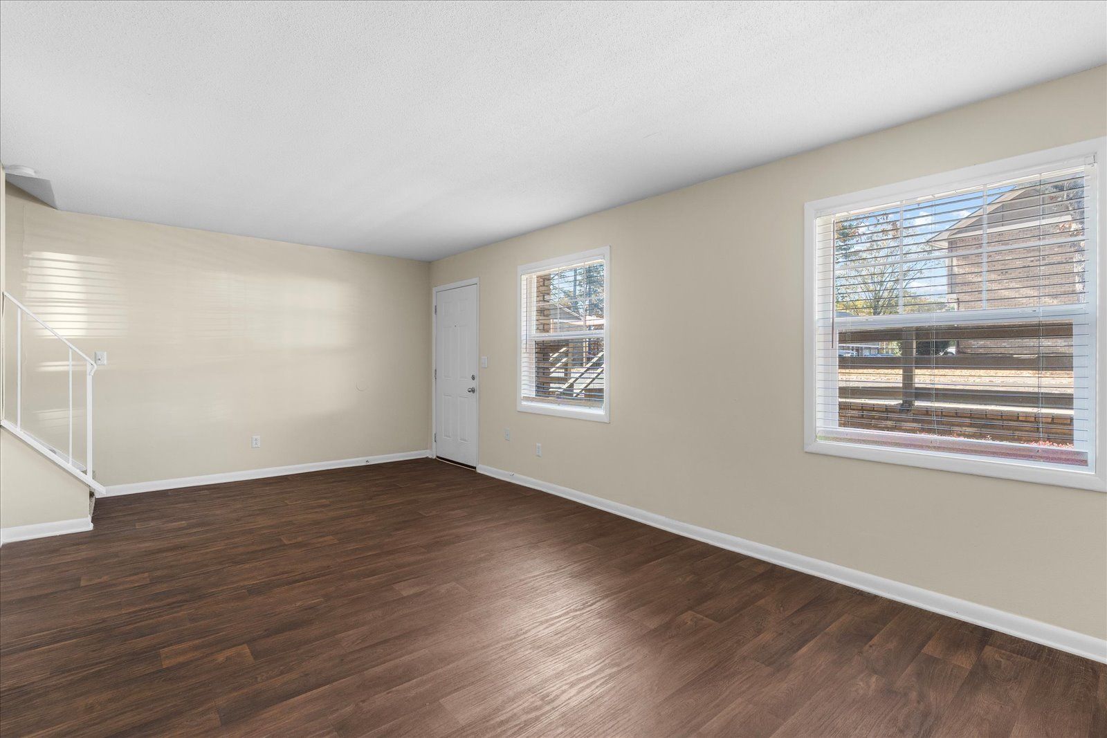 Empty beige room with dark wood floors and two windows letting in daylight.