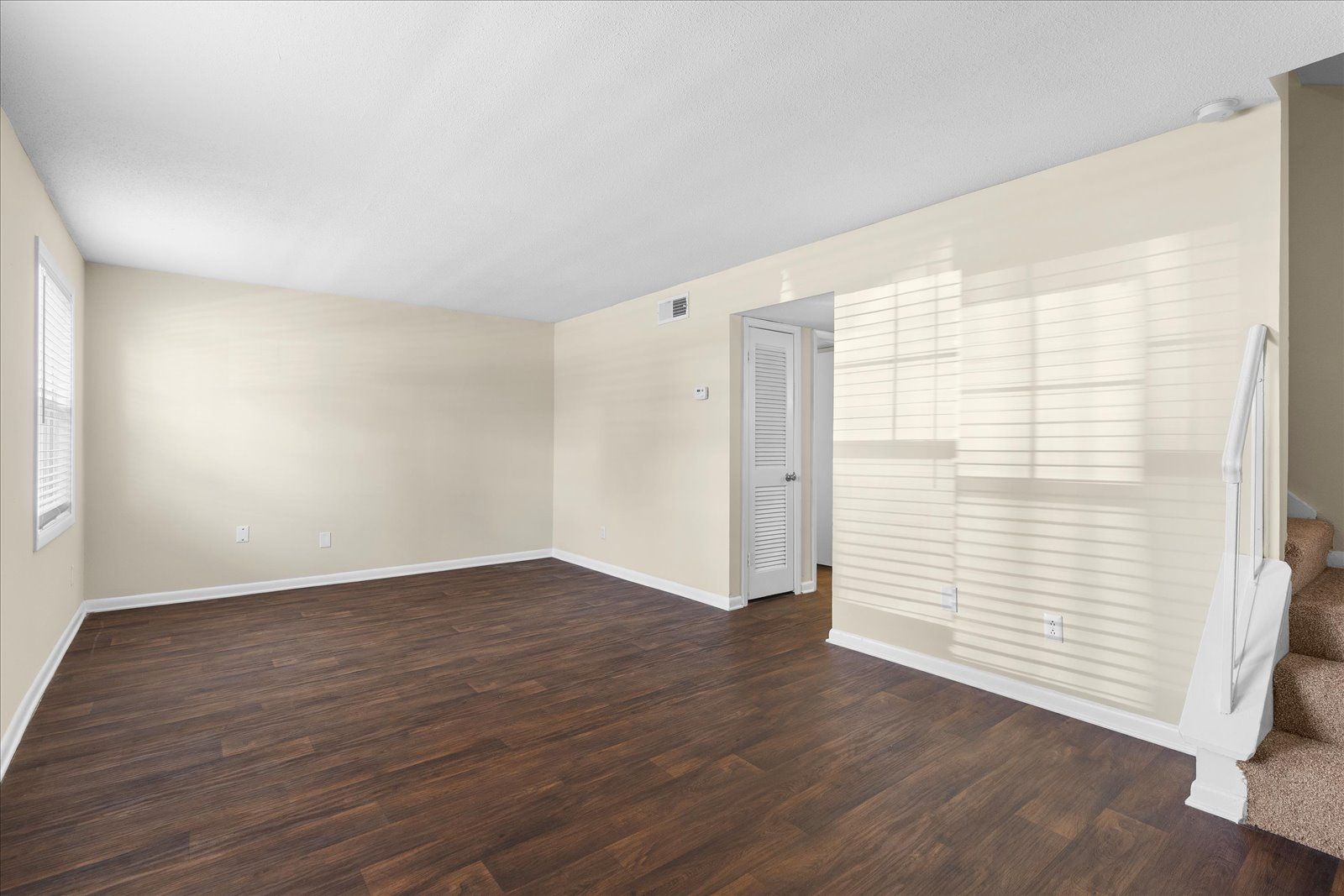 Empty room with dark wood floors, beige walls, and white shutters letting in sunlight